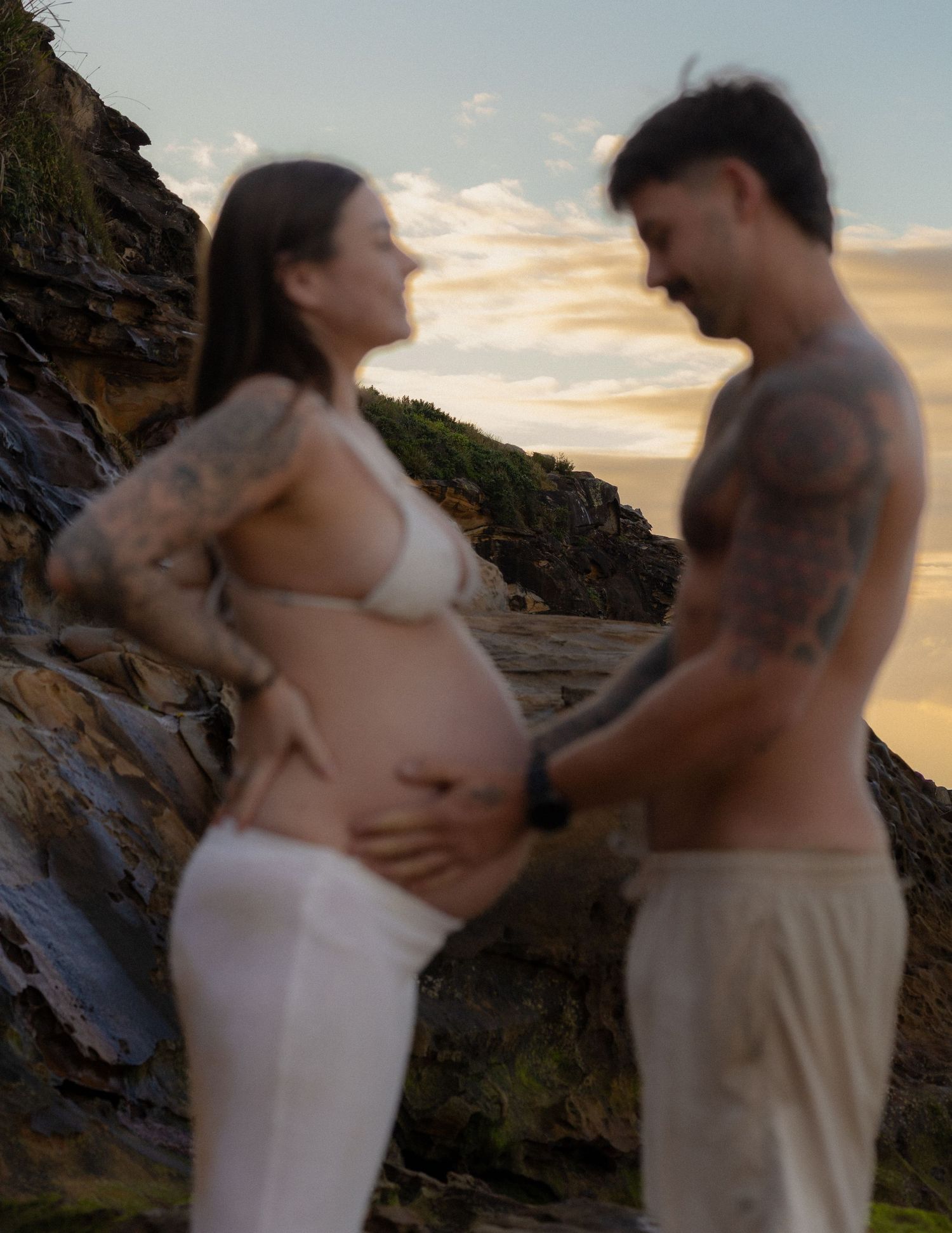 Tattooed couple sharing a tender moment at sunset on rocky coastline, pregnant woman cradling baby bump.