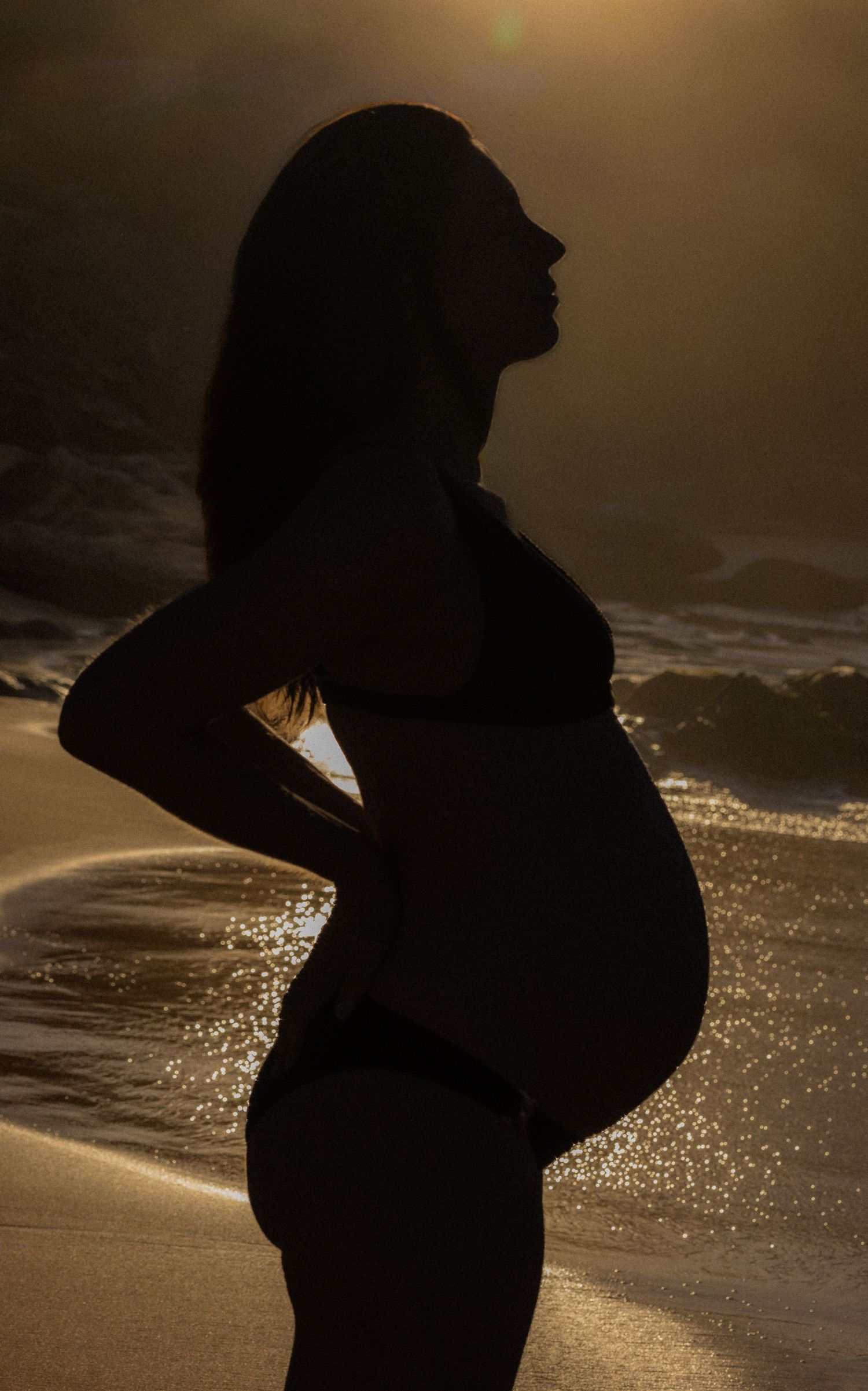 Silhouette of a pregnant woman standing on a beach at golden sunset with ocean waves in the background.