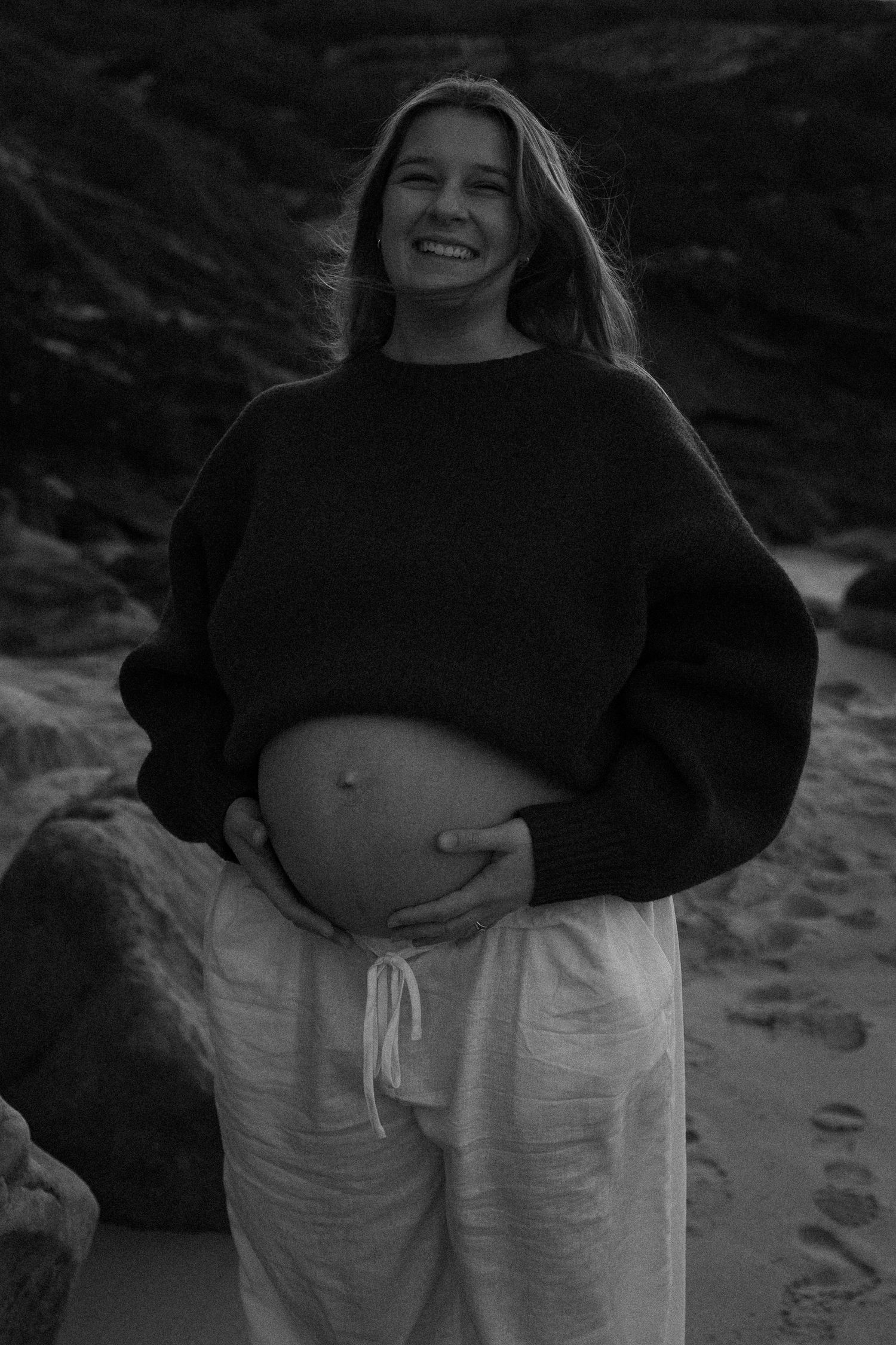 Smiling pregnant woman in black sweater and white linen pants cradling her baby bump on a beach.