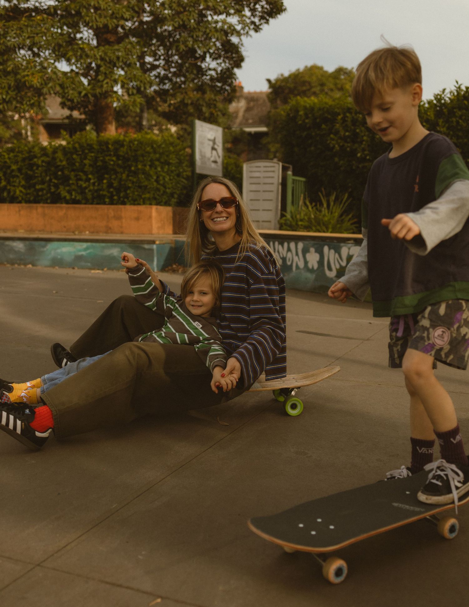 A woman and two children laugh together at a skate park, with kids riding skateboards on a sunny day.