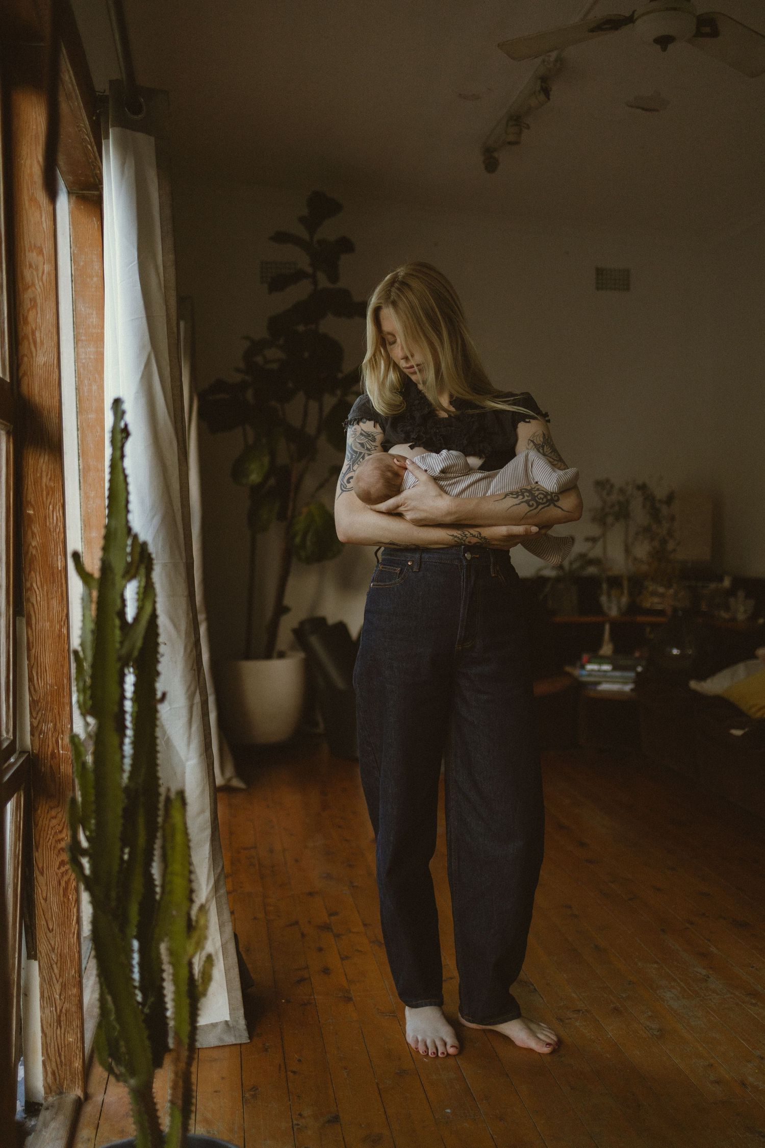 A barefoot mother tenderly holds her newborn baby while standing in a warmly lit, plant-filled living room.