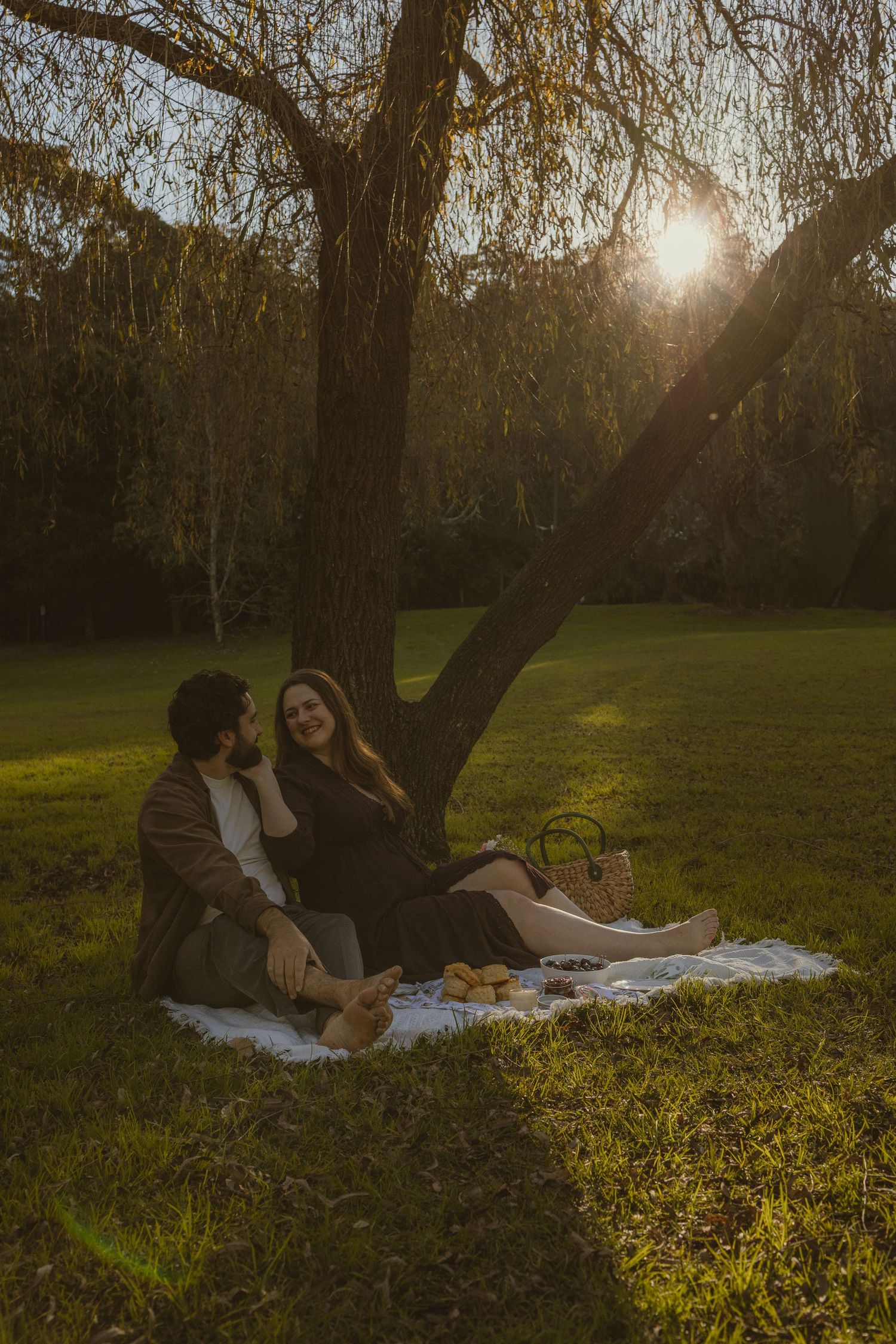 Couple enjoying a romantic sunset picnic on a blanket under a willow tree in a lush green park.