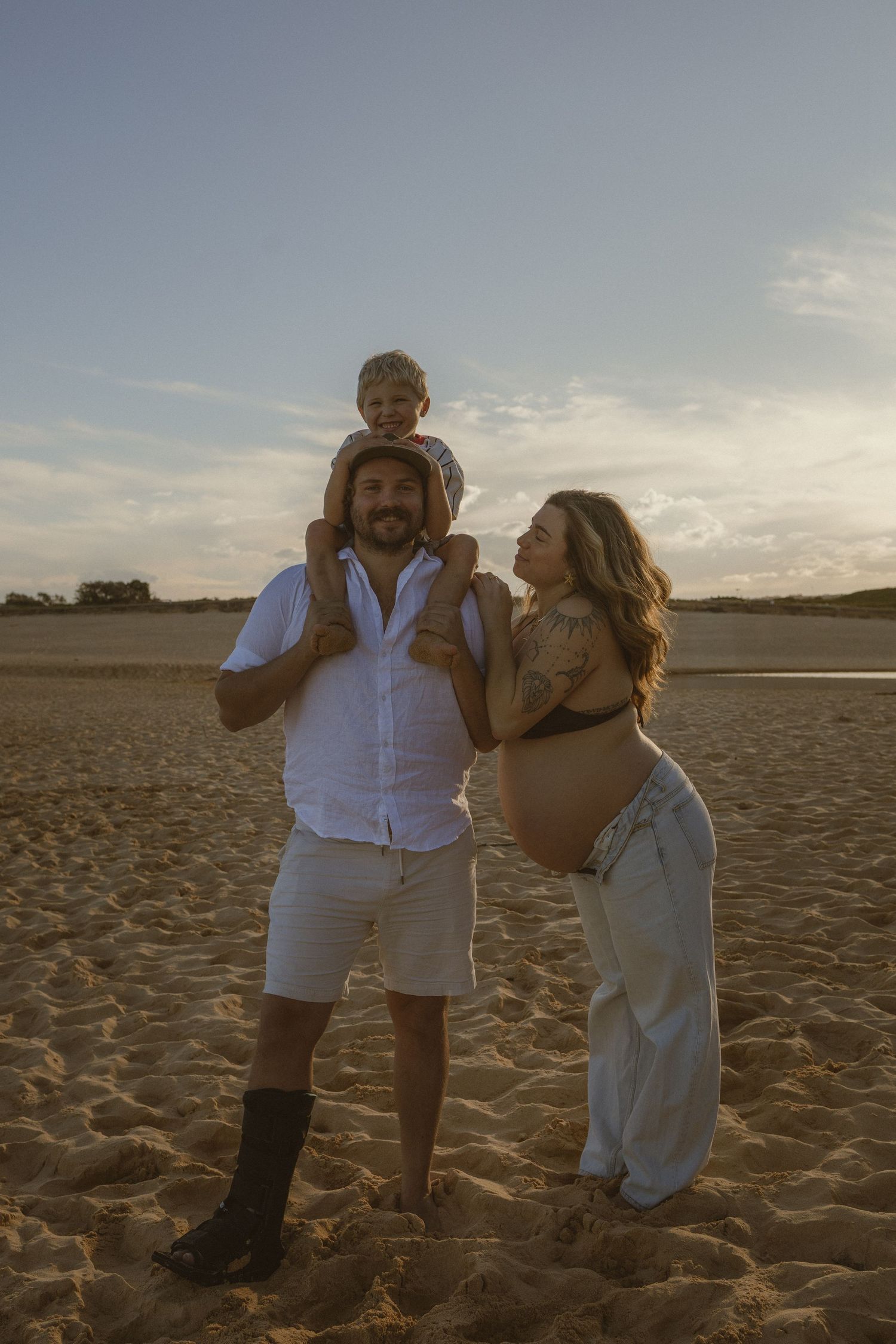 Pregnant woman and man with toddler on shoulders pose on sandy beach at golden hour sunset.