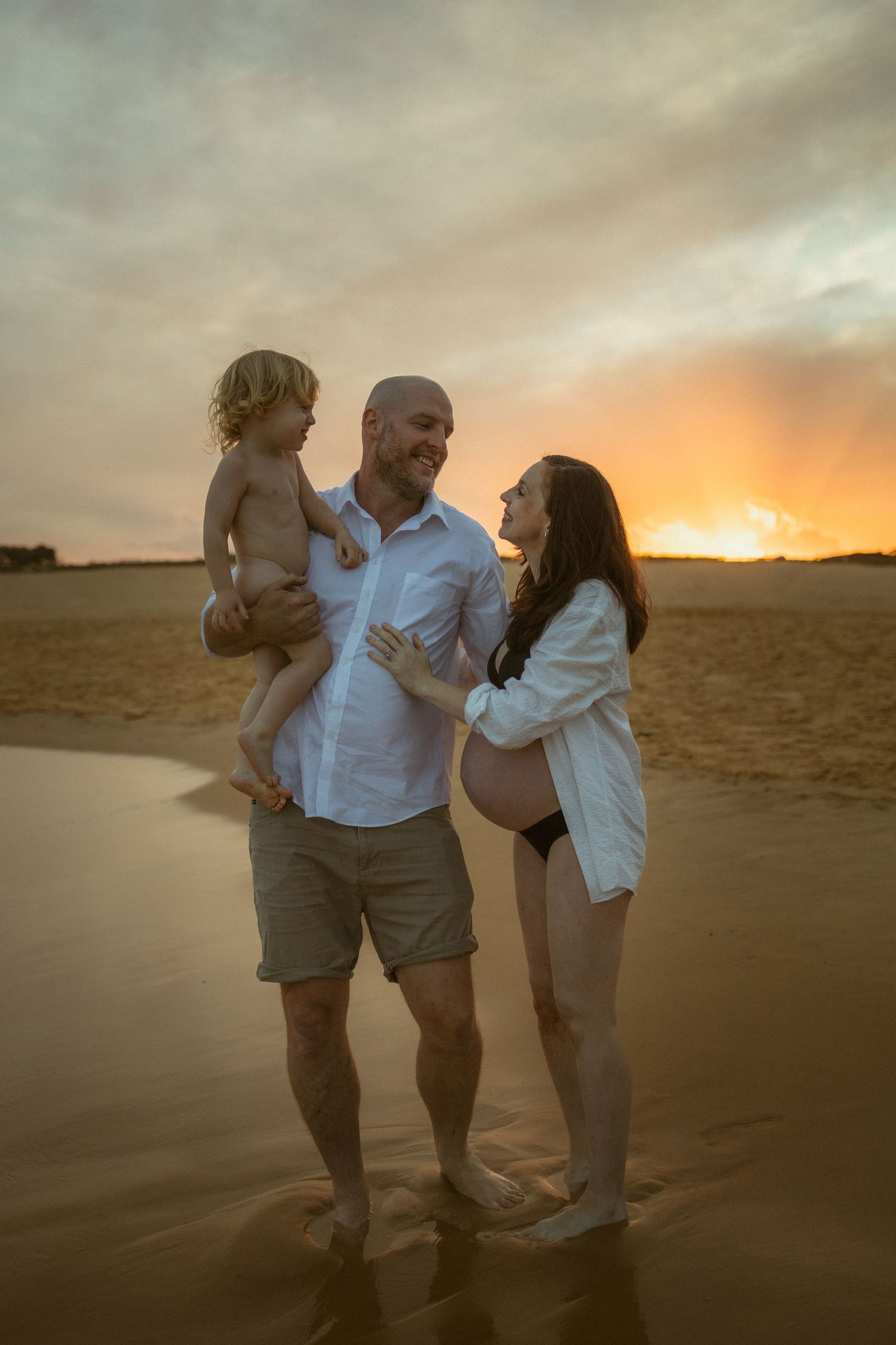Family of three poses on beach at sunset, pregnant mother in bikini and white shirt, father holding toddler son.