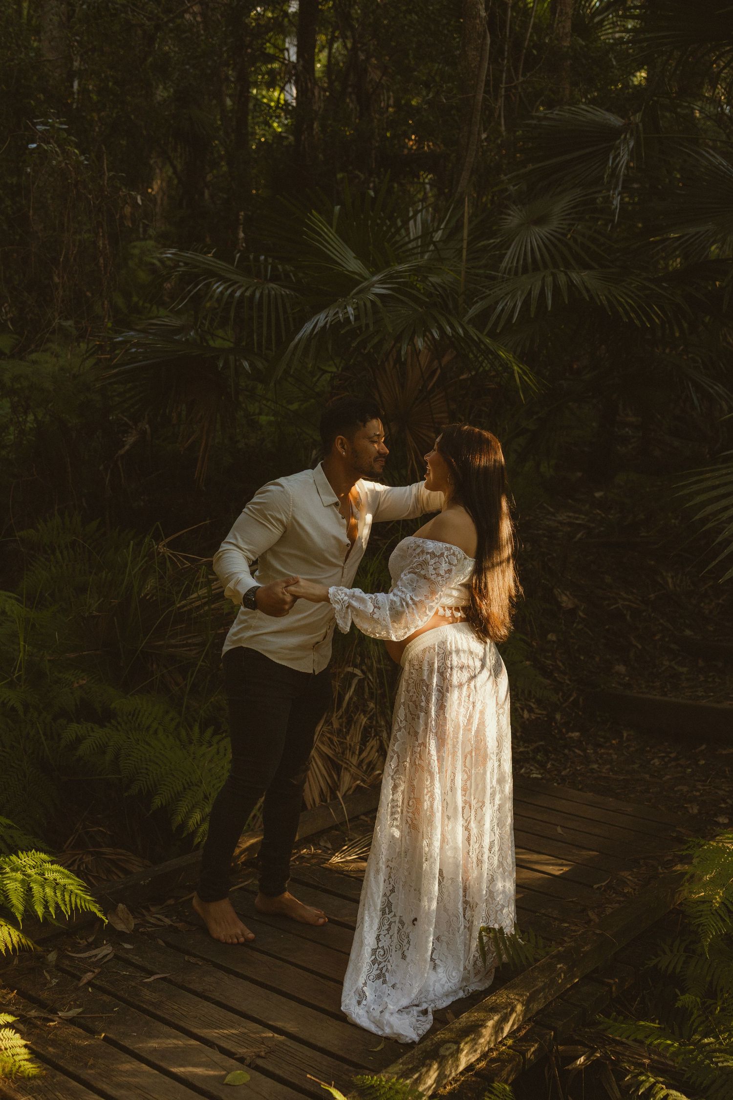 Couple sharing a romantic moment on a wooden bridge surrounded by lush tropical forest, woman in white lace maternity gown.