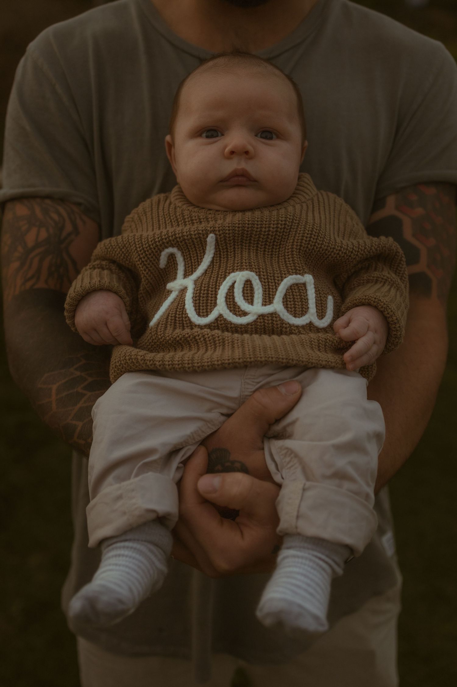 Tattooed father holding newborn baby wearing a brown knit sweater embroidered with the name Koa.