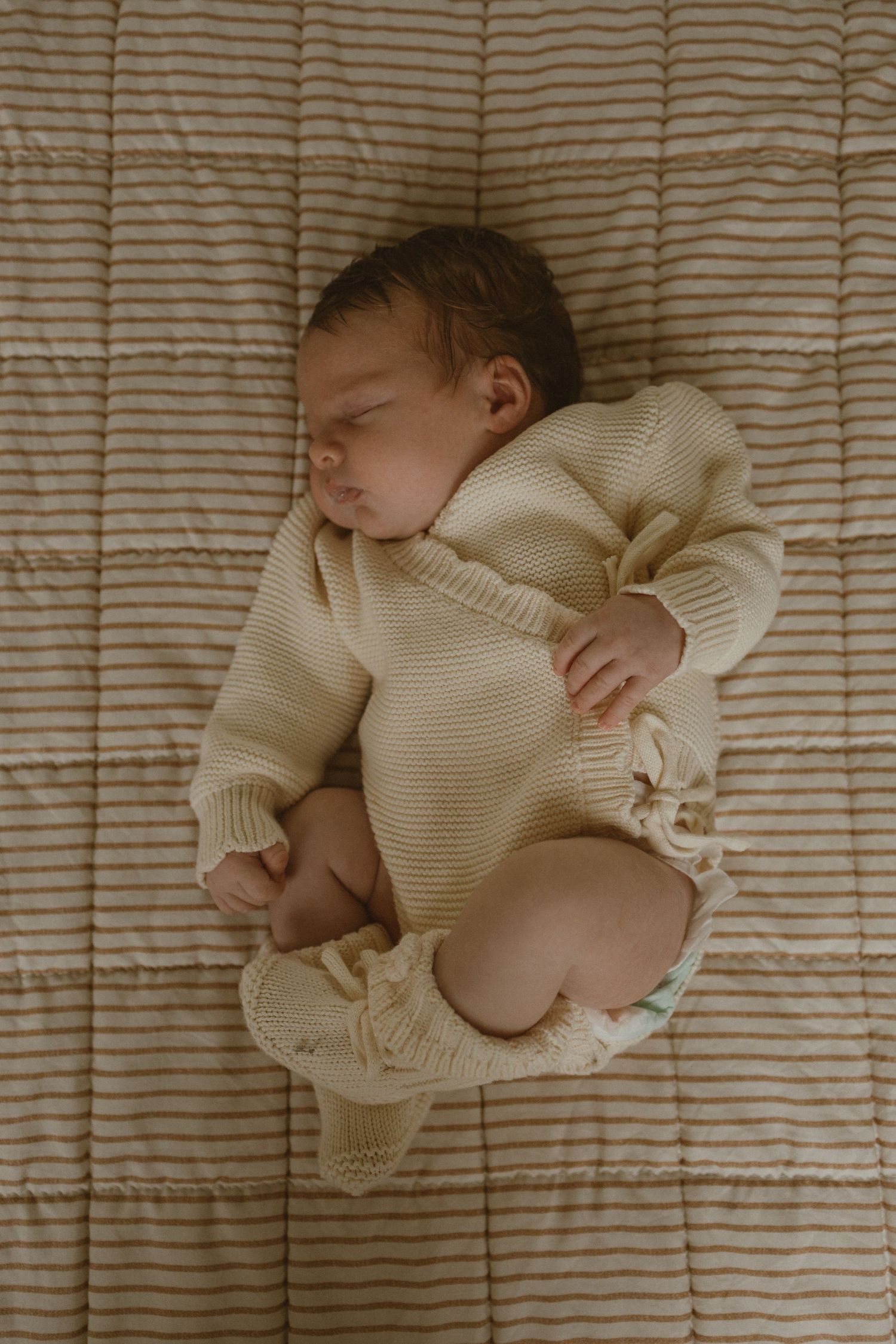 Sleeping newborn baby in cream knit sweater and diaper lying on striped beige quilted blanket.
