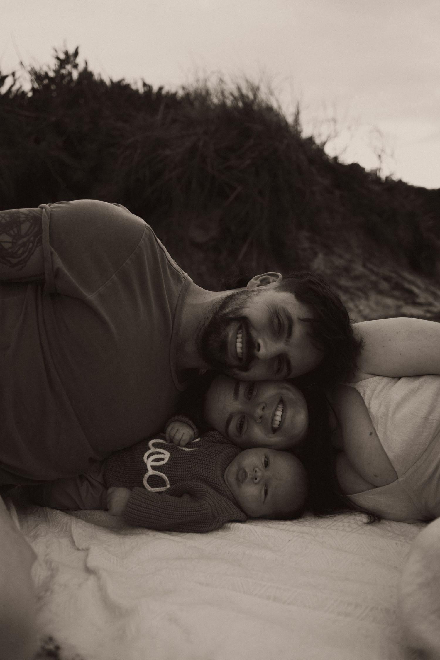 Black and white photo of a bearded father, mother, and newborn baby lying together on a blanket outdoors.