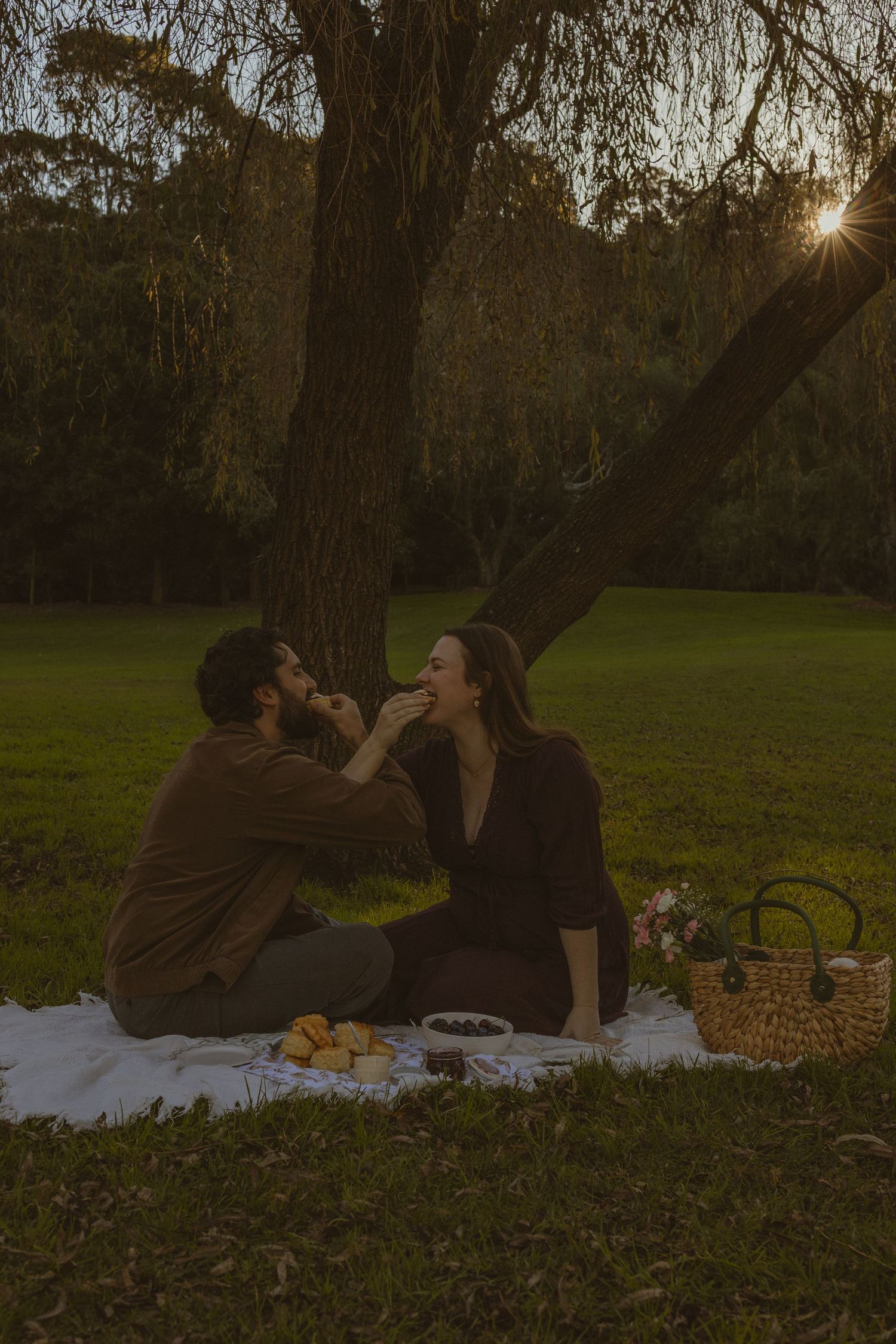 Couple sharing a romantic sunset picnic under a willow tree on a blanket with food and a wicker basket in a park.