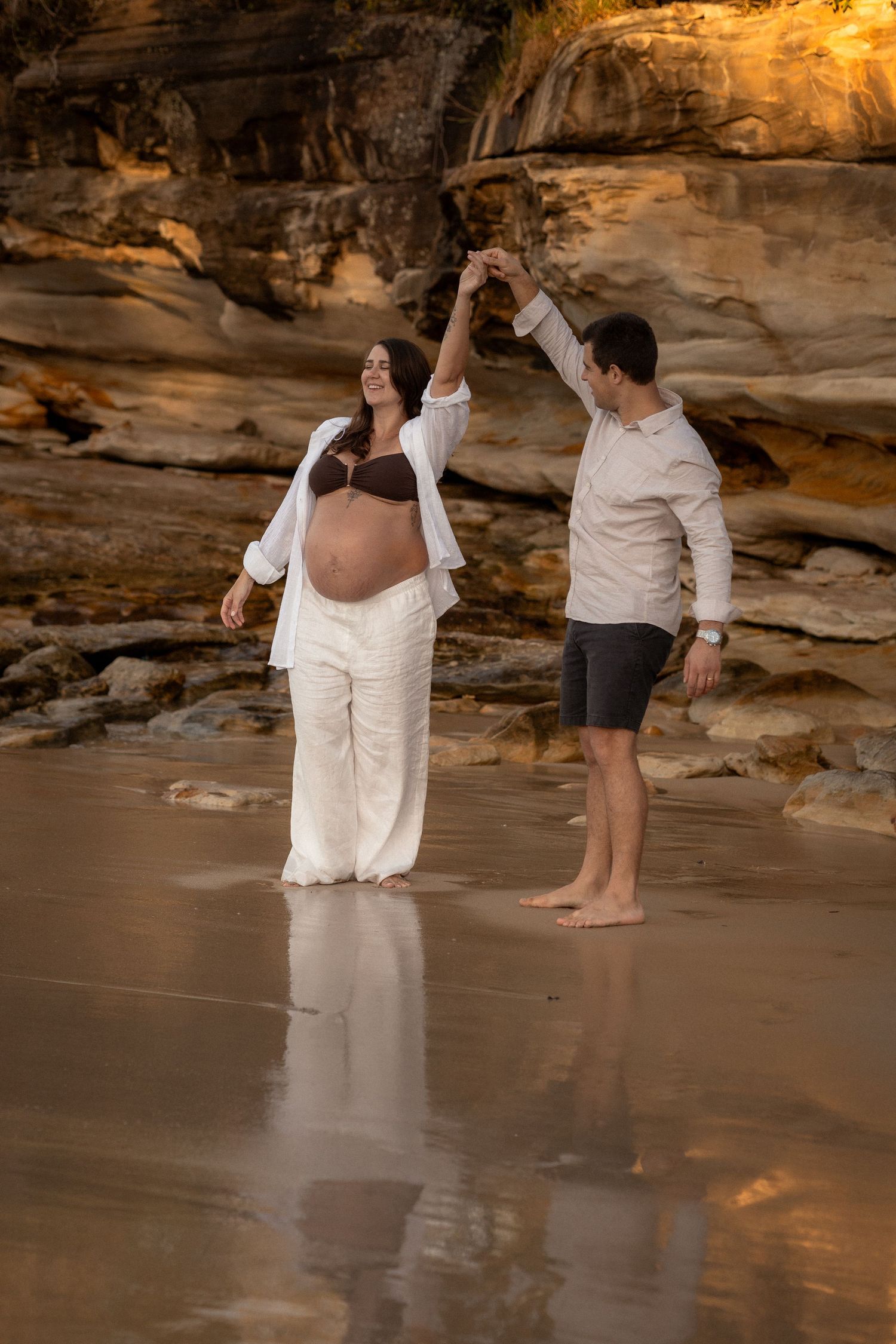 A couple in white attire poses together on a sandy beach at sunset with rocky cliffs in the background.