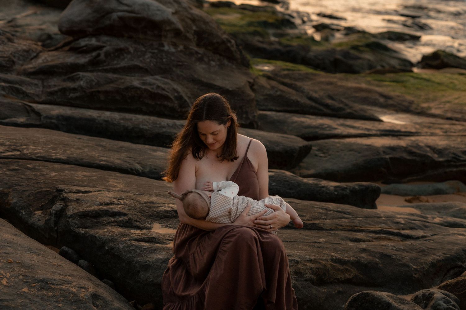 A person in a flowy dress sits on dark rocky terrain at sunset while reading a book.