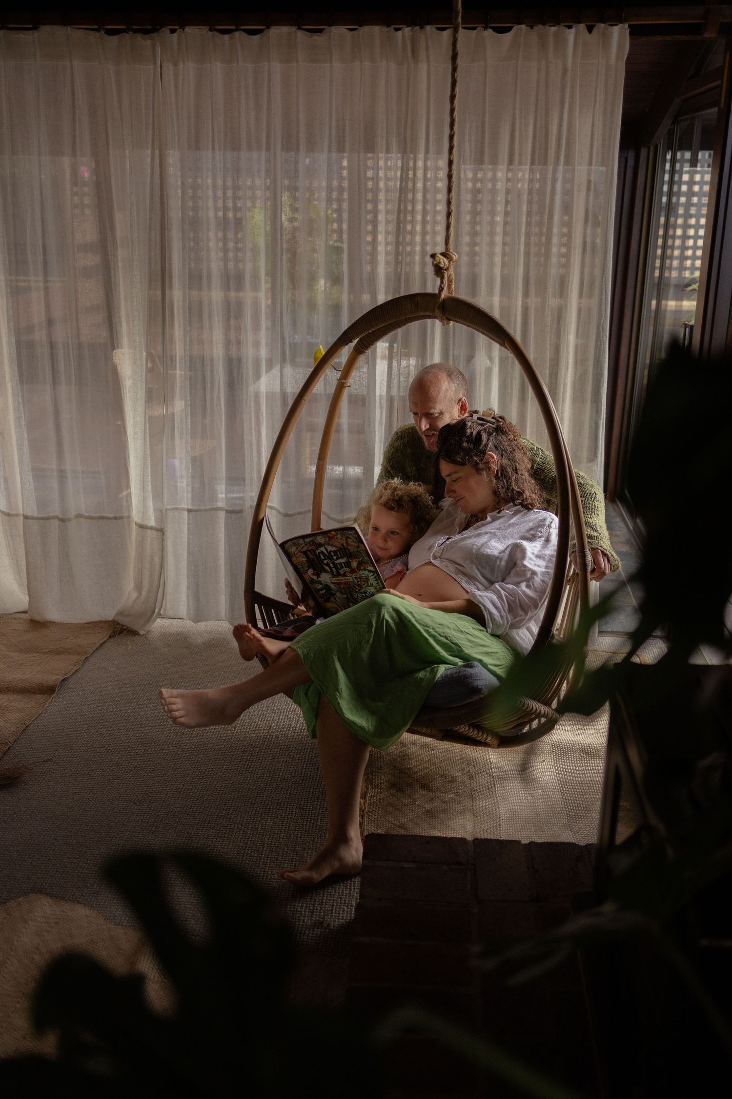Family reading together in a cozy hanging egg chair by sheer curtains in a warm, dimly lit living room.