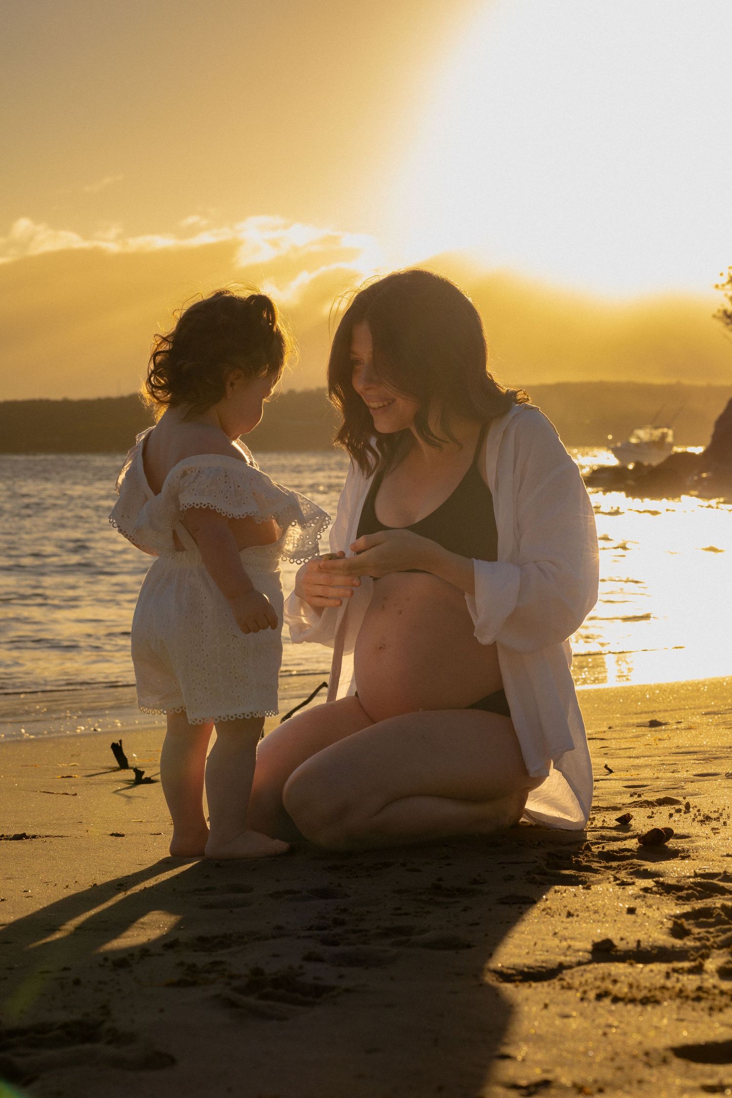 Pregnant woman kneeling on beach at golden sunset with young toddler girl touching her baby bump.
