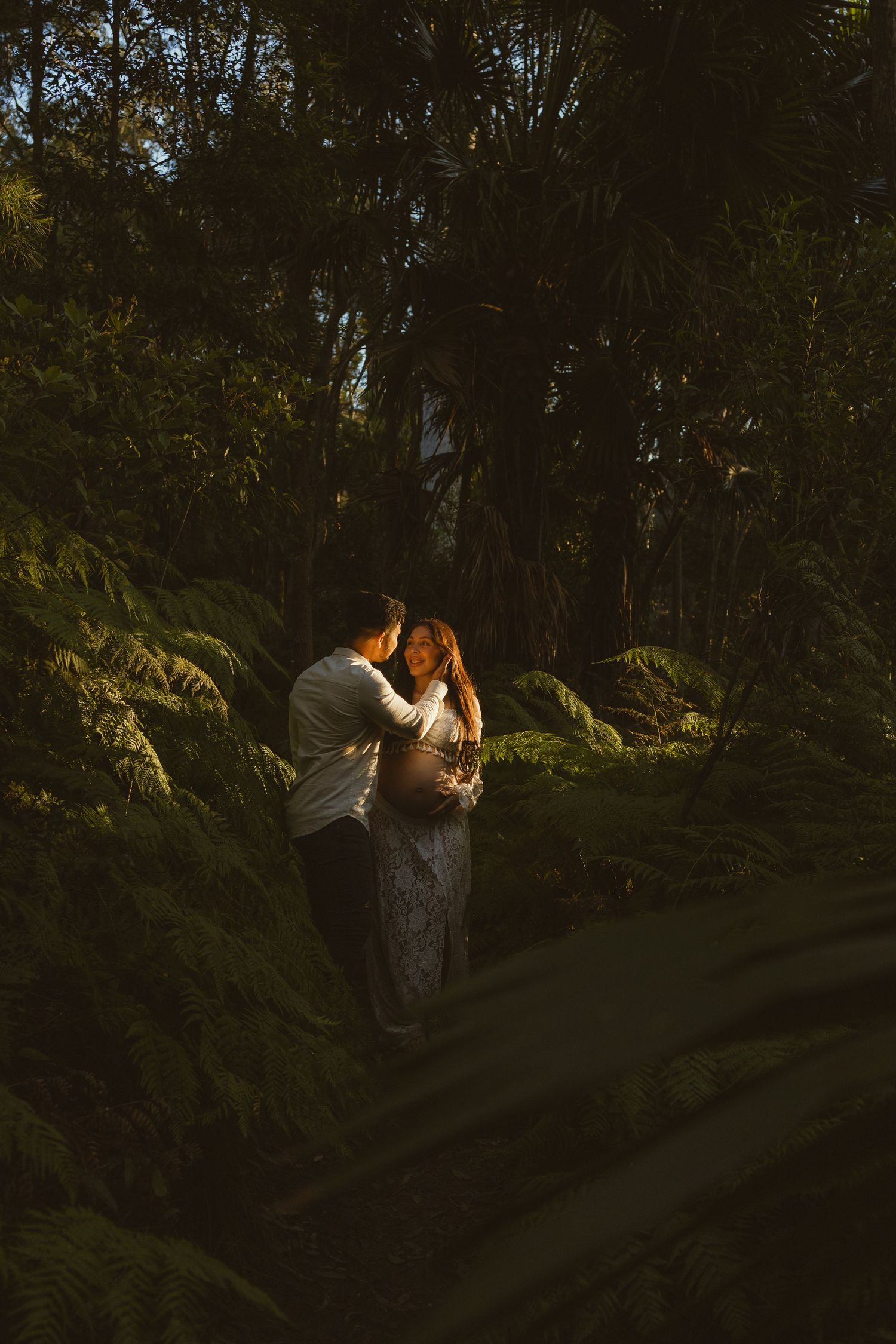 Couple embracing tenderly in dark lush forest, golden light illuminating pregnant woman in white lace gown.