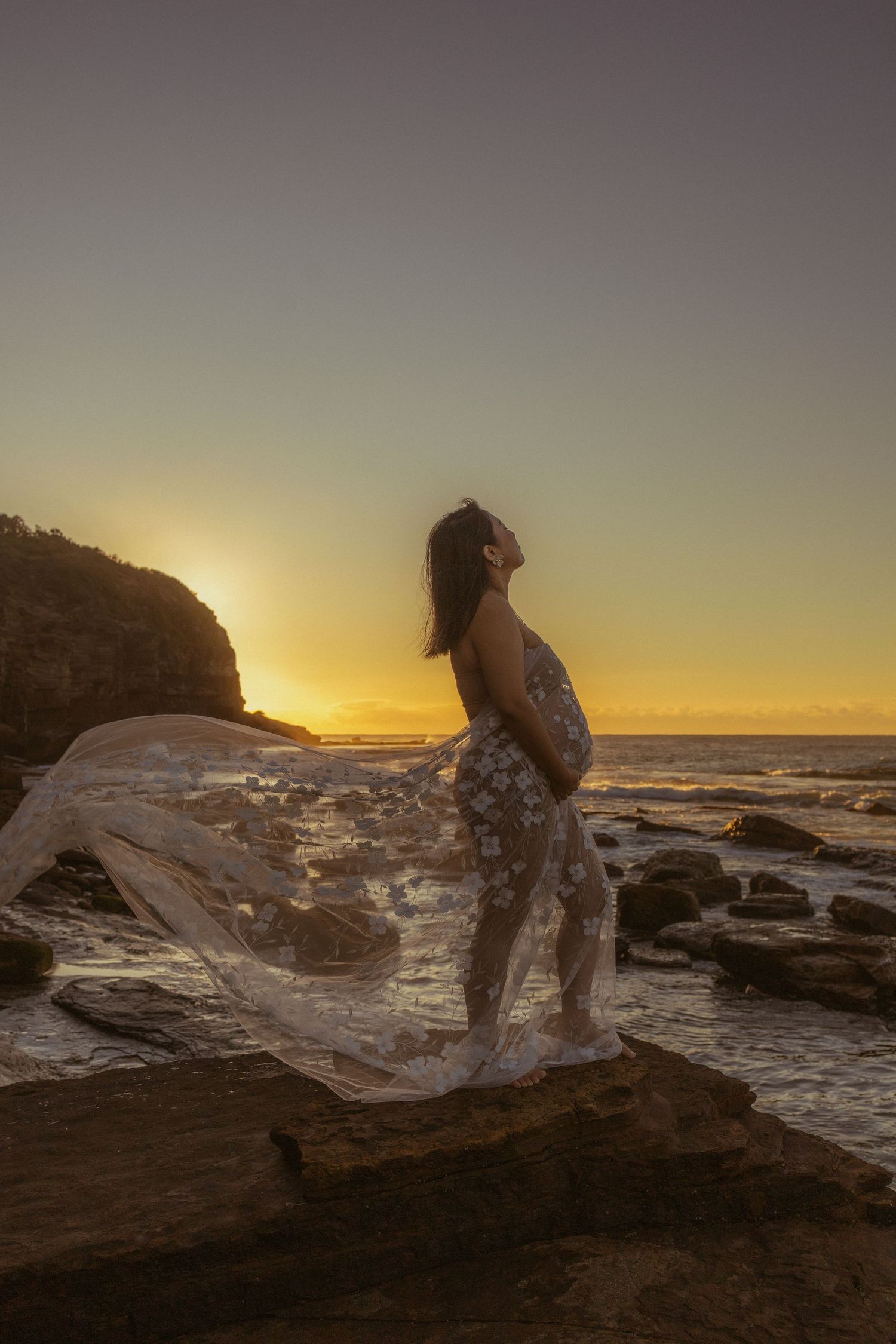 Pregnant woman in flowing lace dress standing on rocks at golden sunset by the ocean.