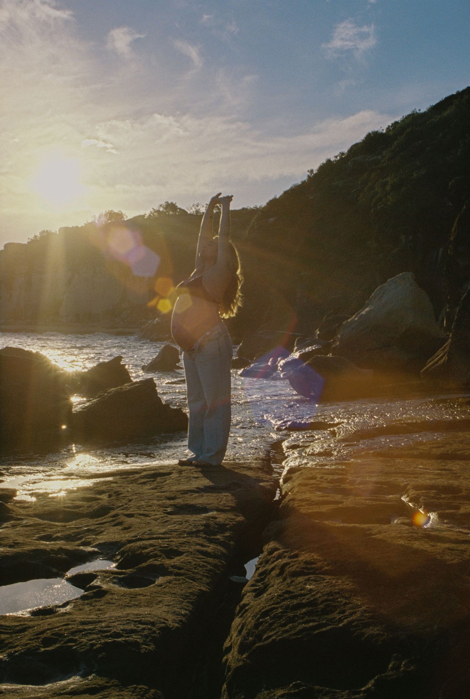 Woman stretches arms up on rocky coastal shore during stunning golden sunset.