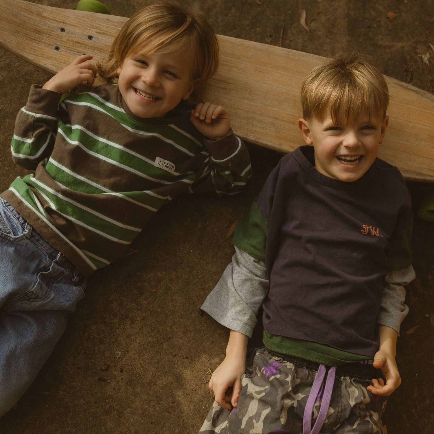 Two smiling young boys lying on the ground, one holding a green skateboard, wearing casual striped and navy tops.