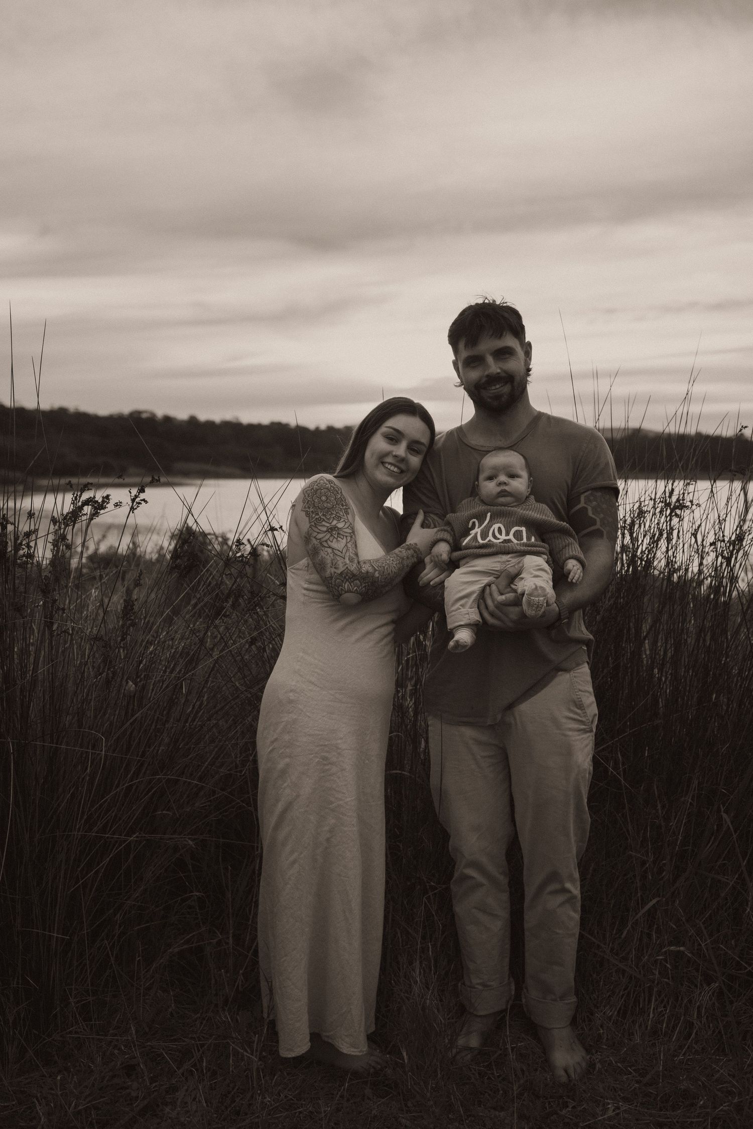 Sepia-toned family portrait of a couple with their baby standing in tall grass near a lake under cloudy skies.