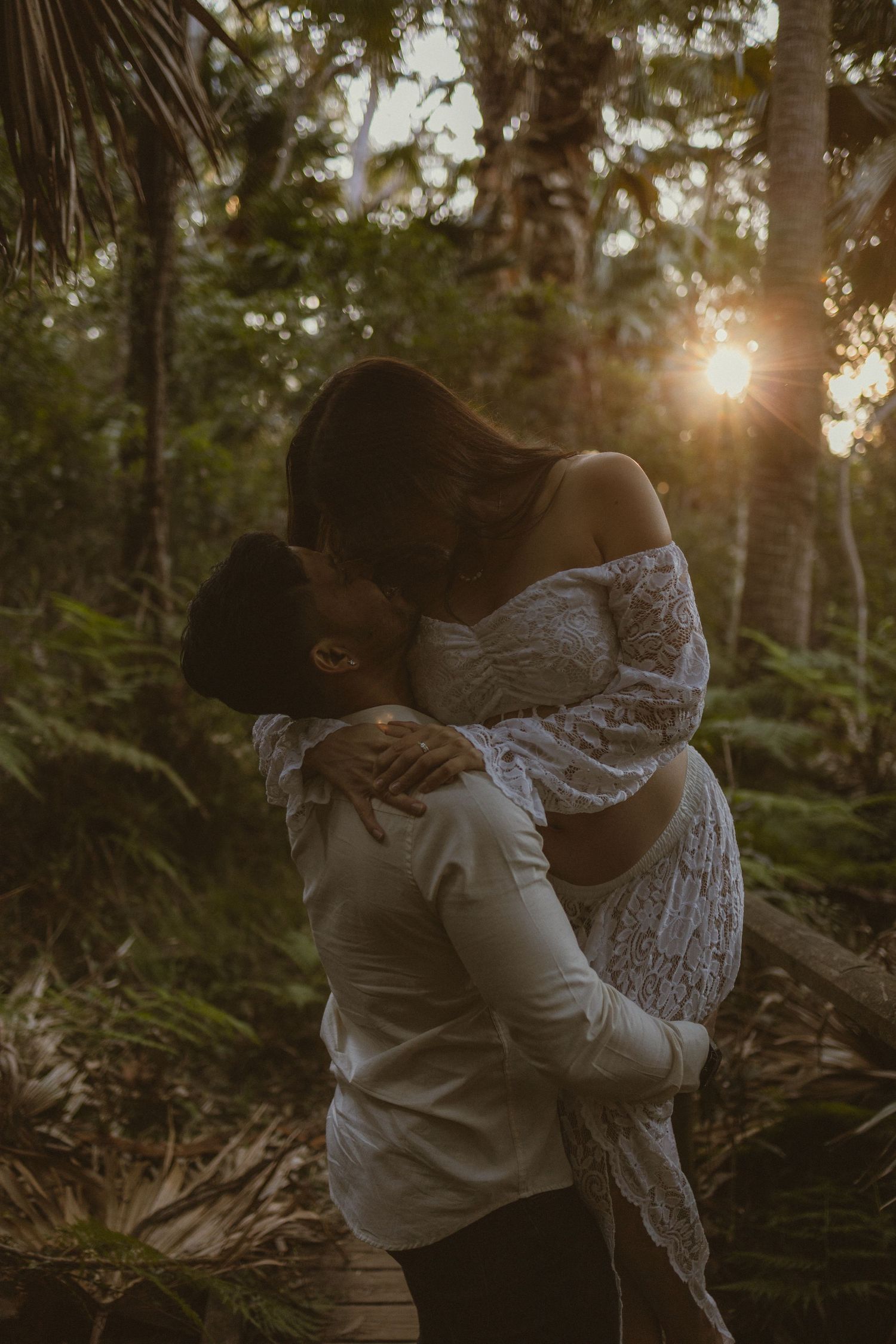 Couple sharing a romantic kiss in a lush forest at golden sunset, man lifting woman in white lace maternity dress.