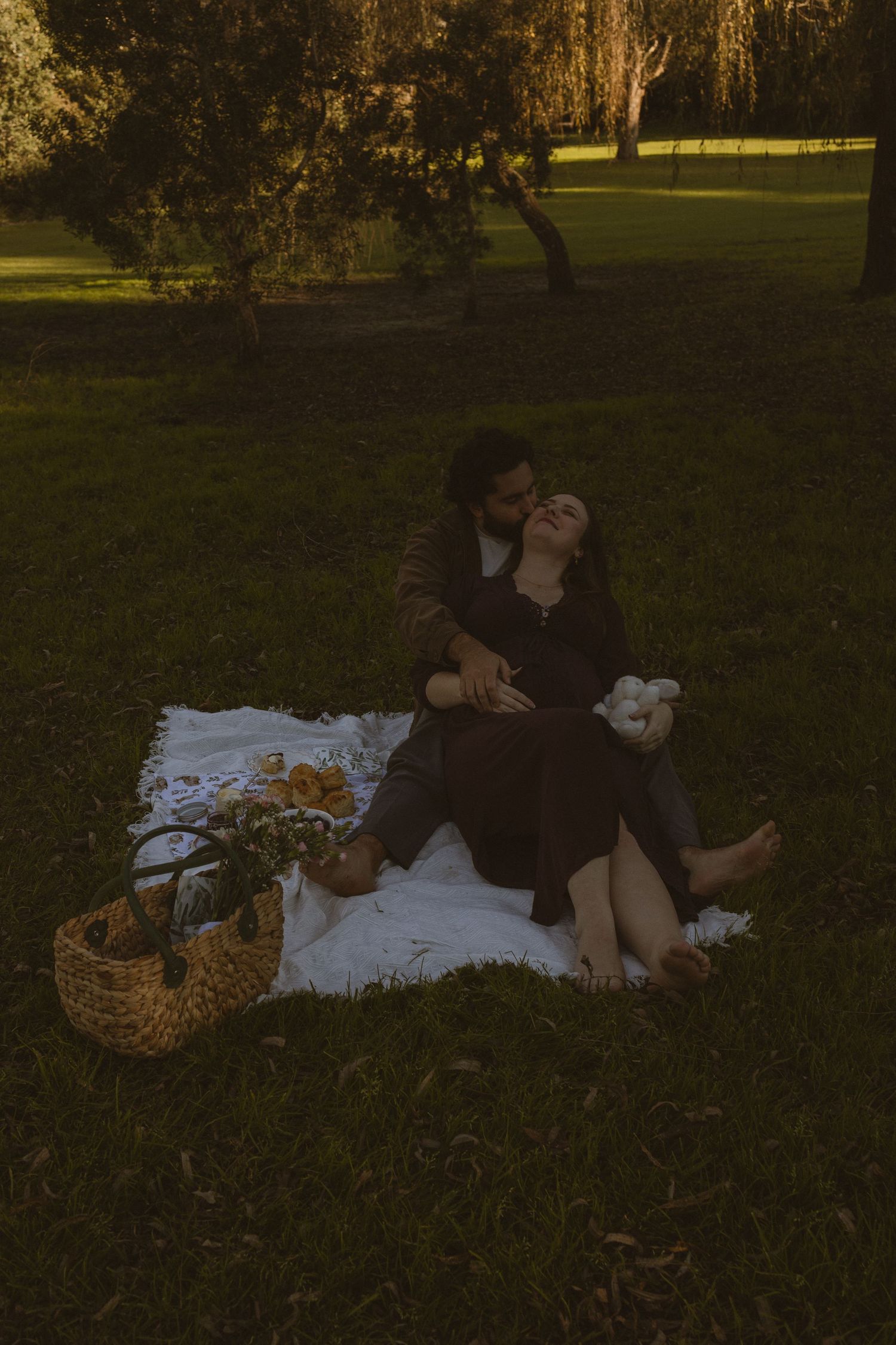 Couple enjoying a romantic picnic on a blanket in a lush green park at dusk, with a wicker basket and flowers nearby.