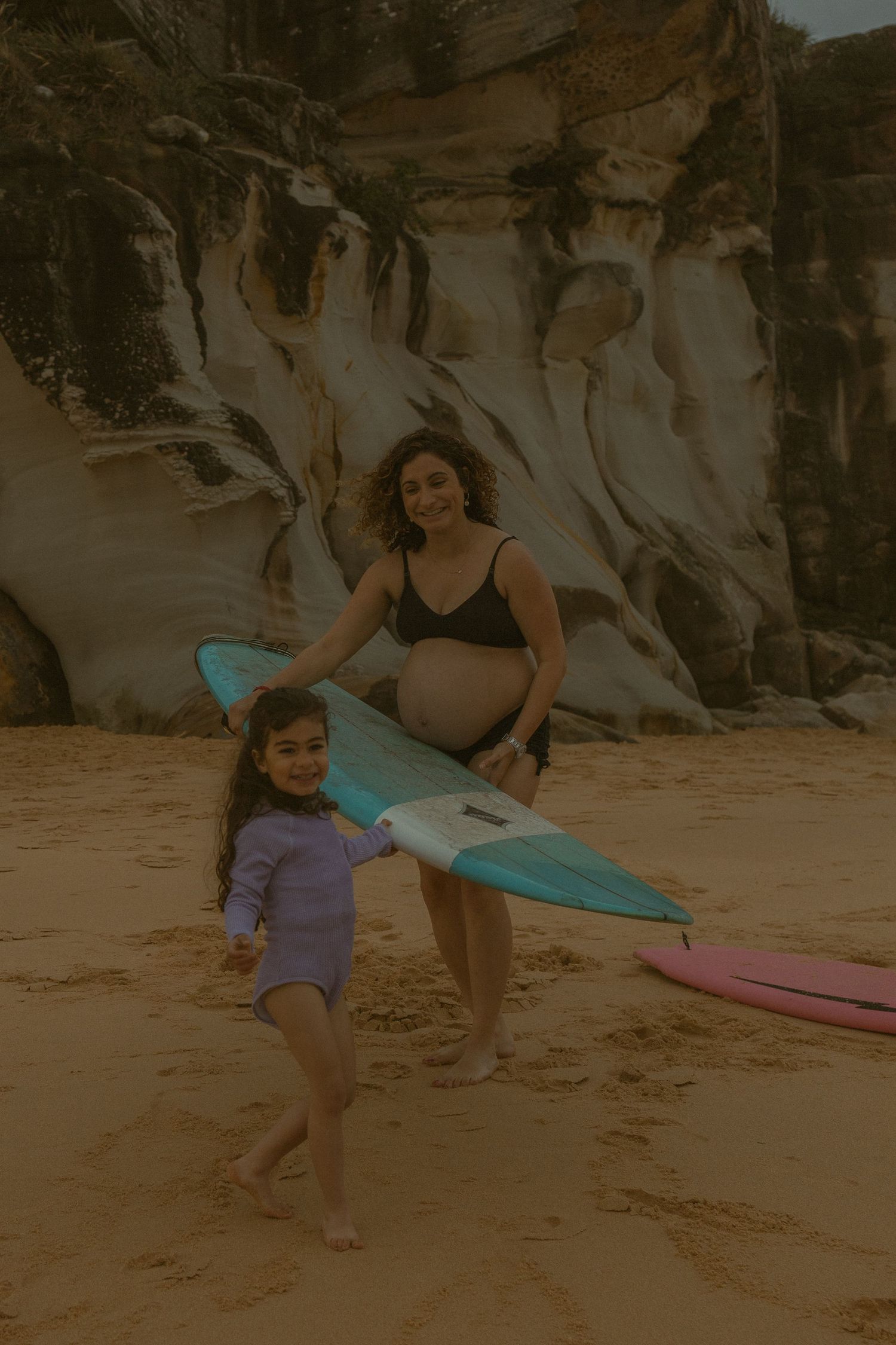 Pregnant woman in black bikini holds blue surfboard with young girl on sandy beach near rocky cliffs.