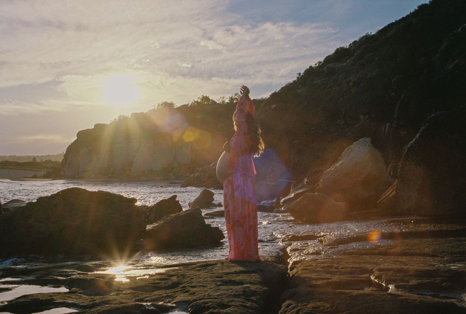 Woman in pink dress standing on rocky beach at sunset with golden light and ocean waves.