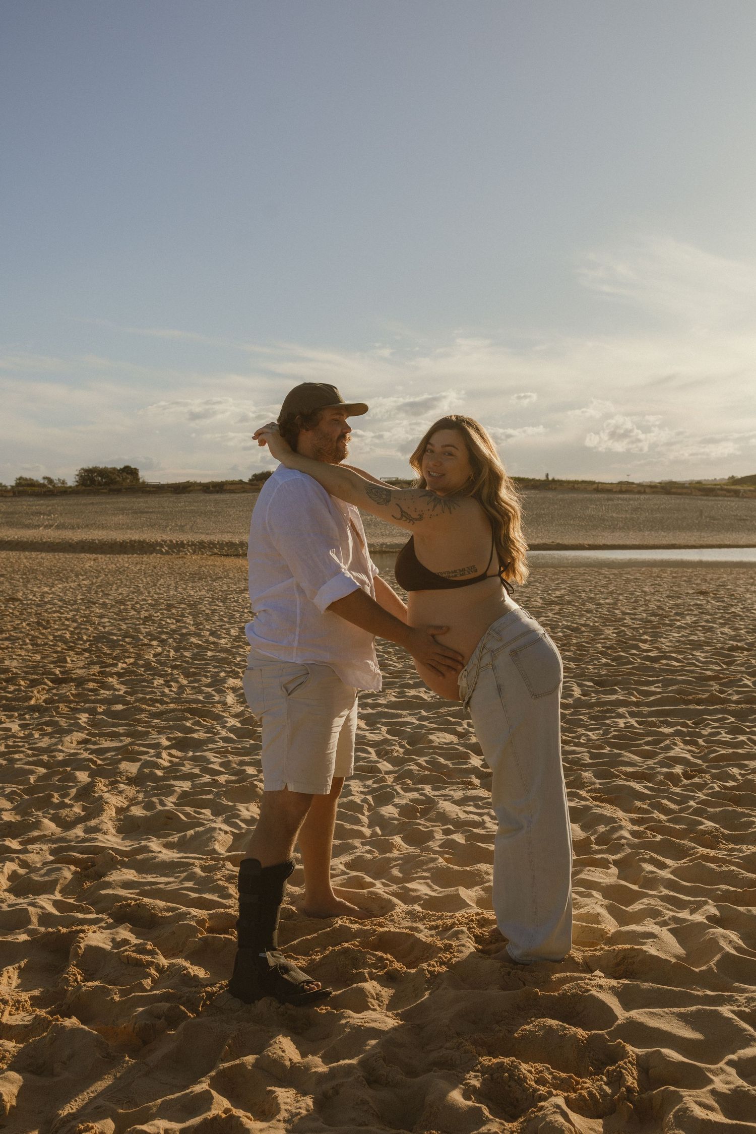Couple embracing on sandy beach at golden hour, woman in bikini top and white pants, man in white shirt and cap.
