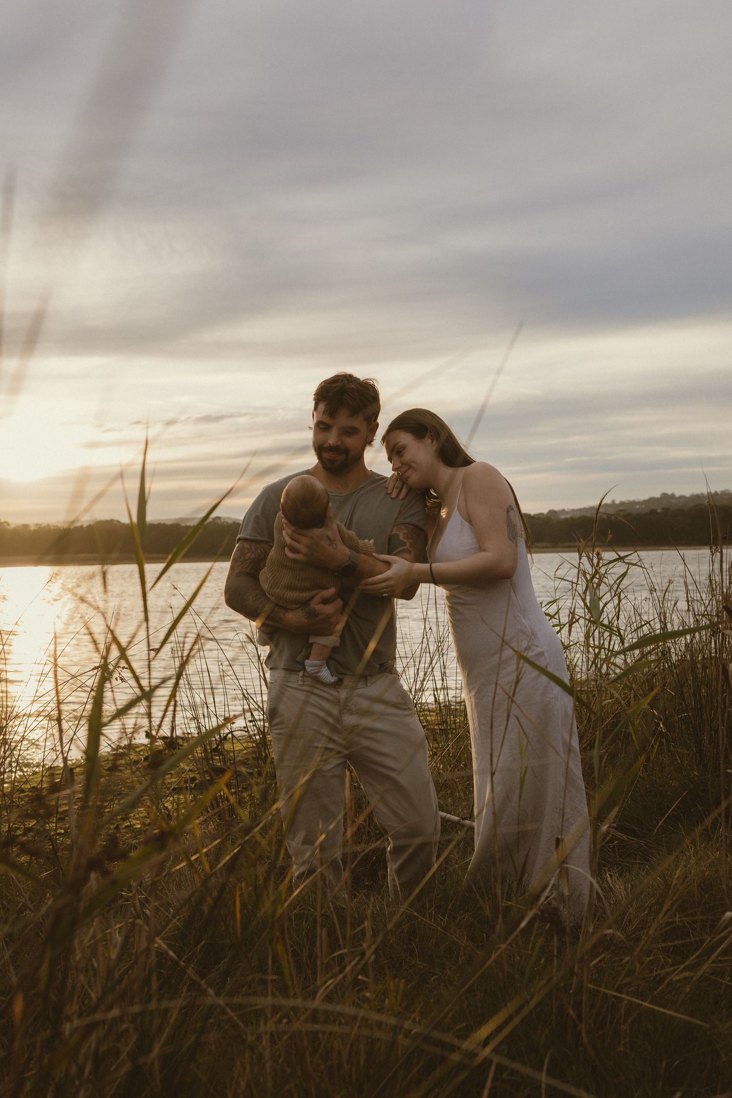 A couple stands in tall grass by a lake at sunset, tenderly holding their newborn baby in a warm golden light.