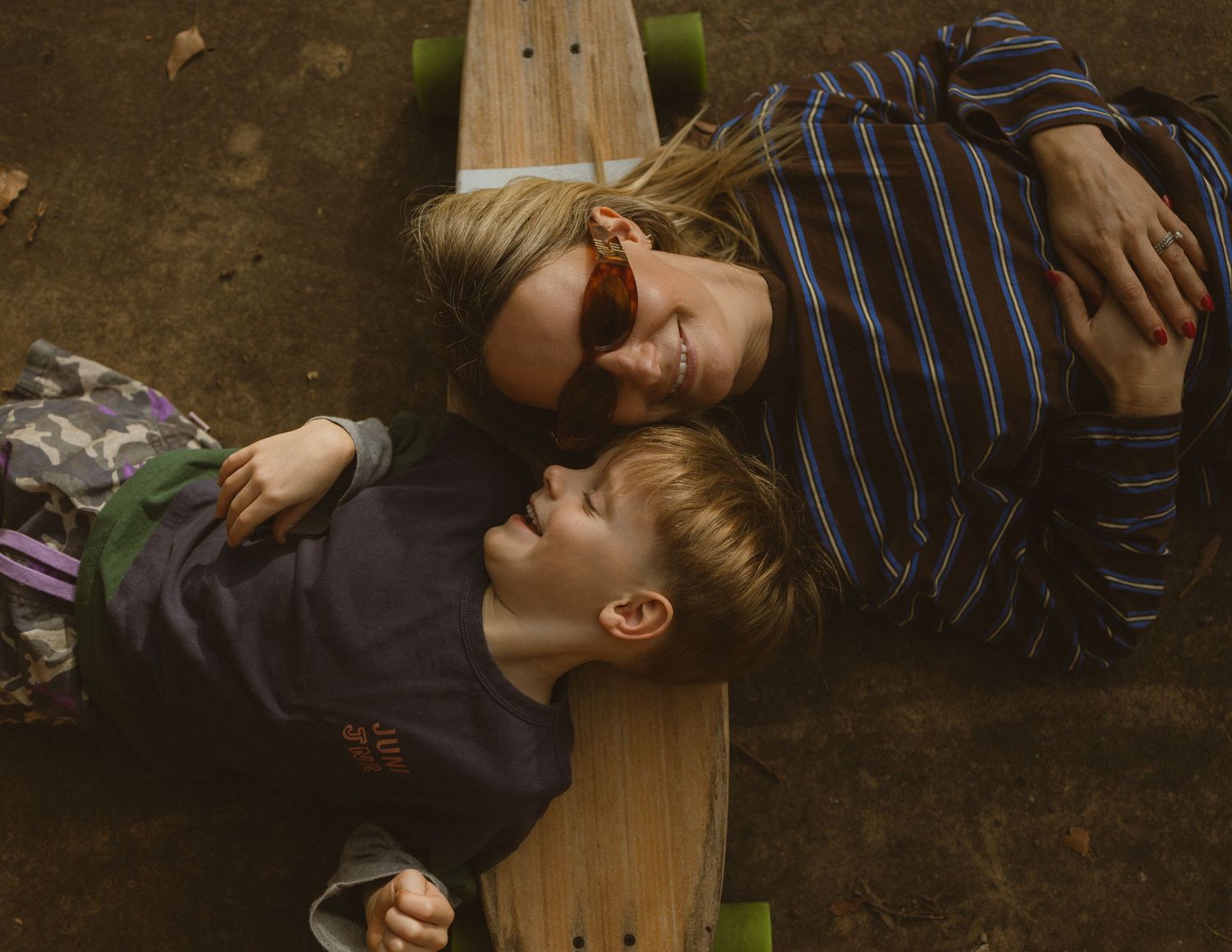 Two children lying on a skateboard outdoors, smiling at each other in a candid moment.
