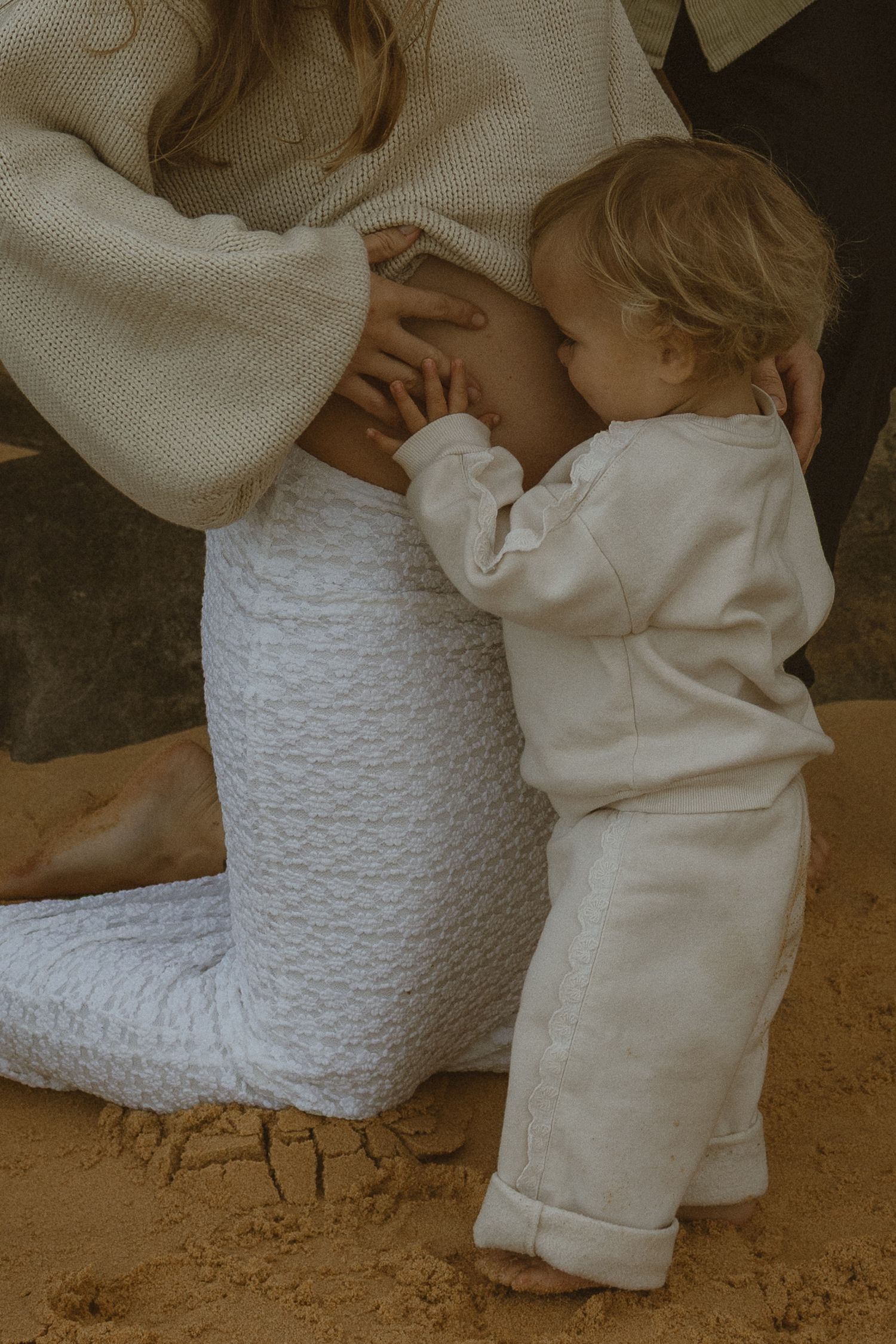 Toddler in white outfit standing on sand, hugging a kneeling pregnant woman in cream knit sweater and white textured pants.