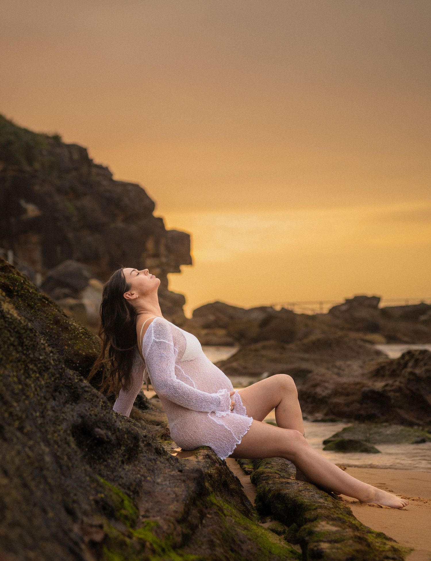 A figure in a white dress reclines on rocky coastal cliffs during a dramatic golden sunset.