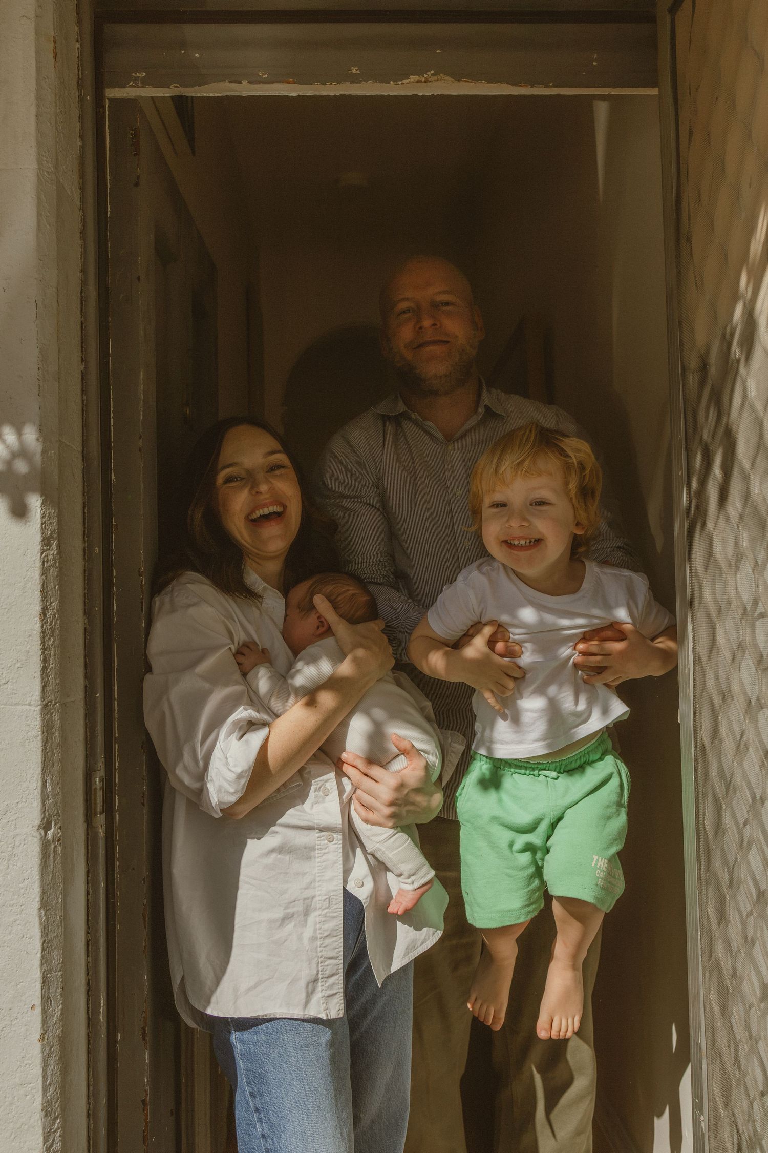Happy family of four posing in sunny doorway, mother holding newborn, father lifting smiling toddler in green shorts.