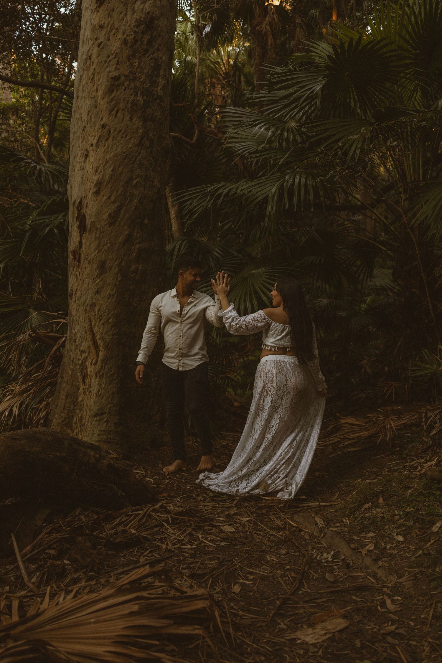 Couple dancing together in a lush tropical forest, woman in white lace skirt reaching toward man in white shirt.