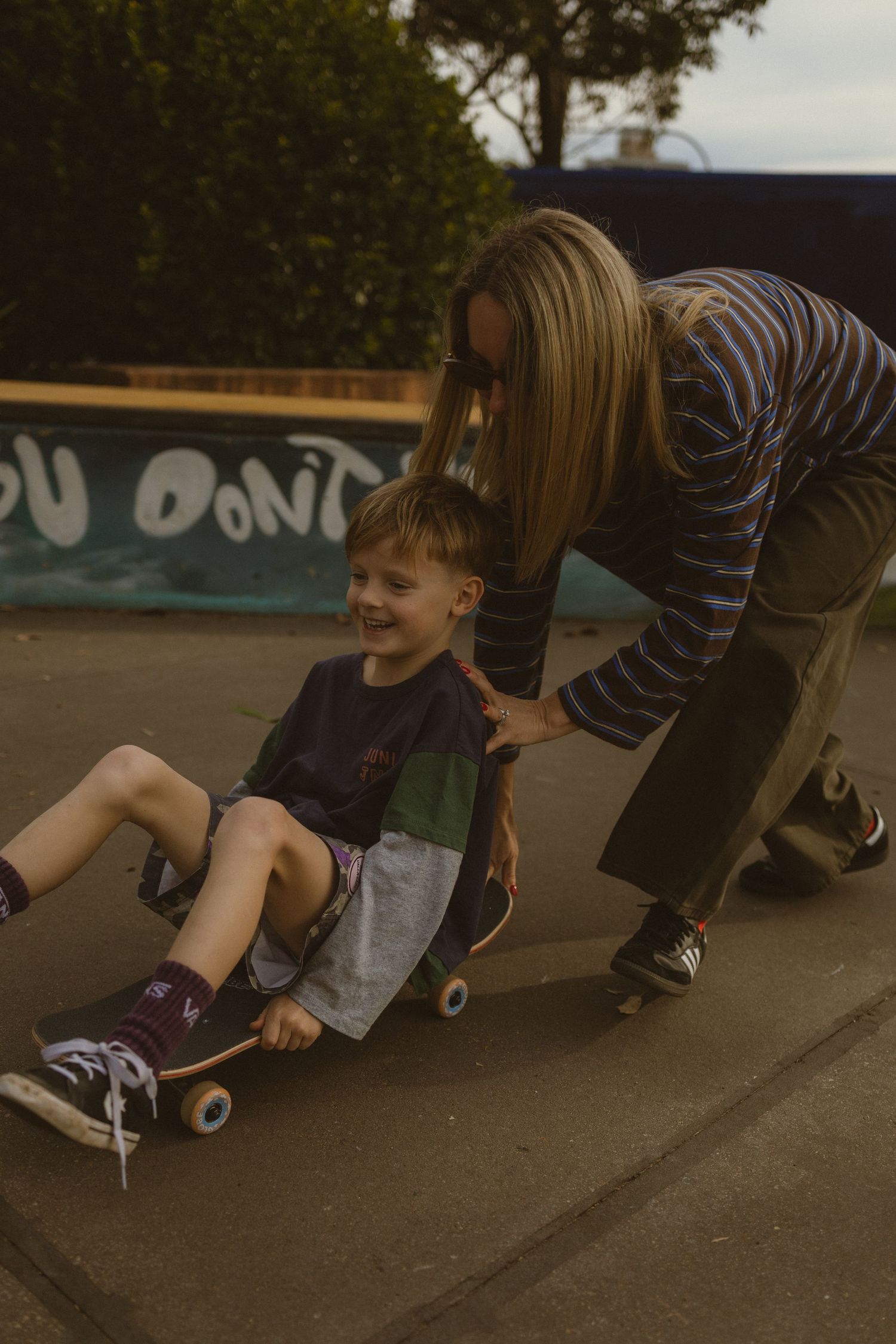 A woman helps a smiling young boy ride a skateboard at an outdoor skate park.