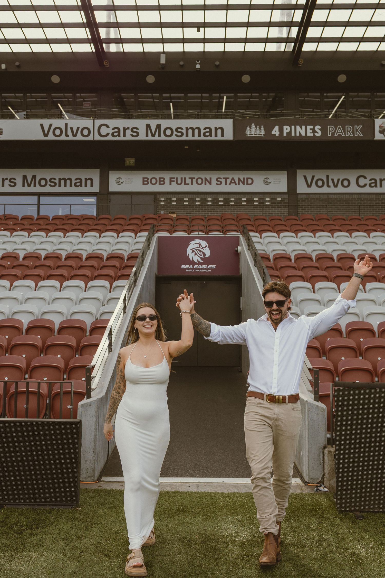 Couple celebrating at 4 Pines Park stadium, Bob Fulton Stand, raising hands joyfully in white outfits.