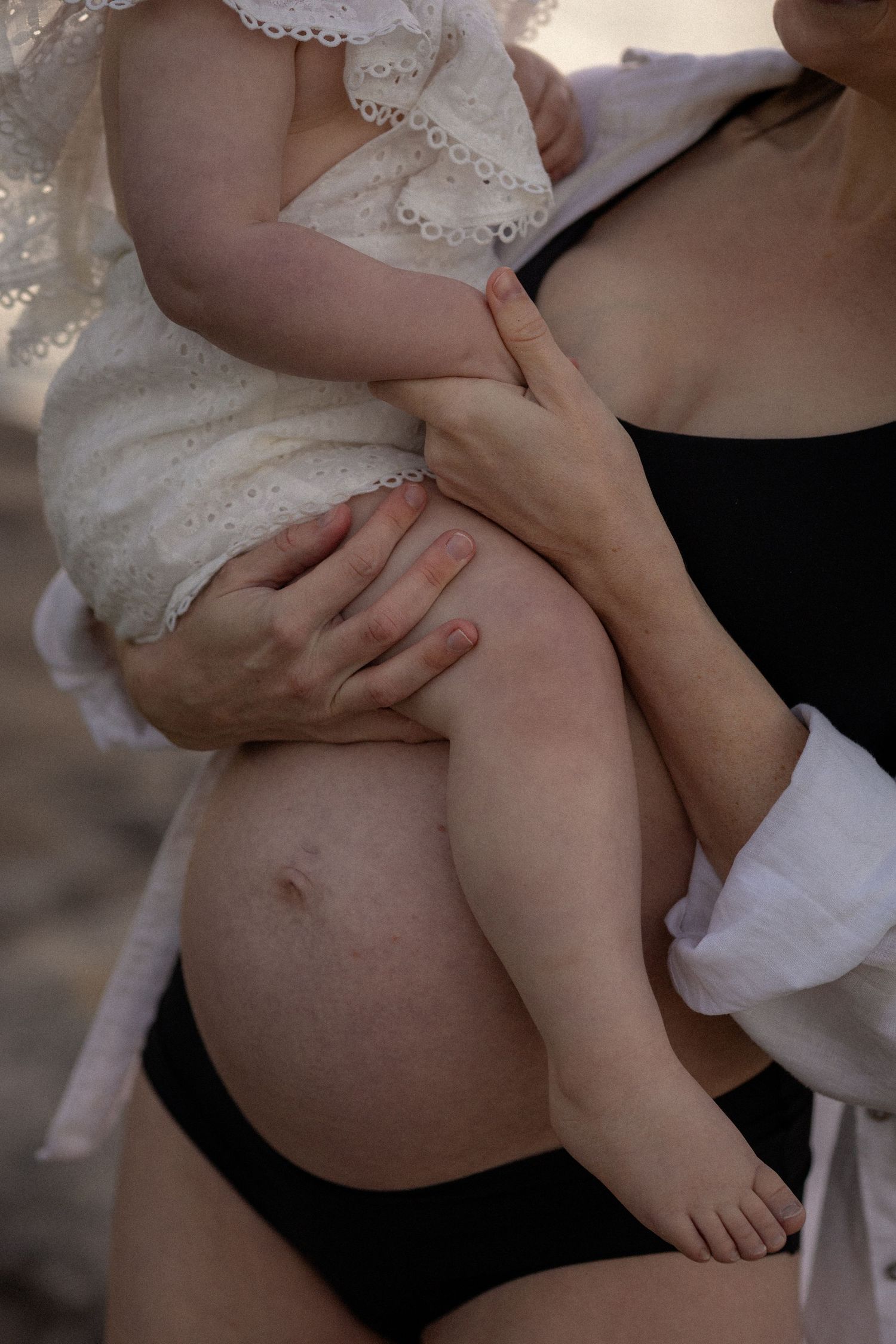 Pregnant woman in black bikini bottom holding a baby dressed in white lace outfit, showcasing maternity and motherhood.