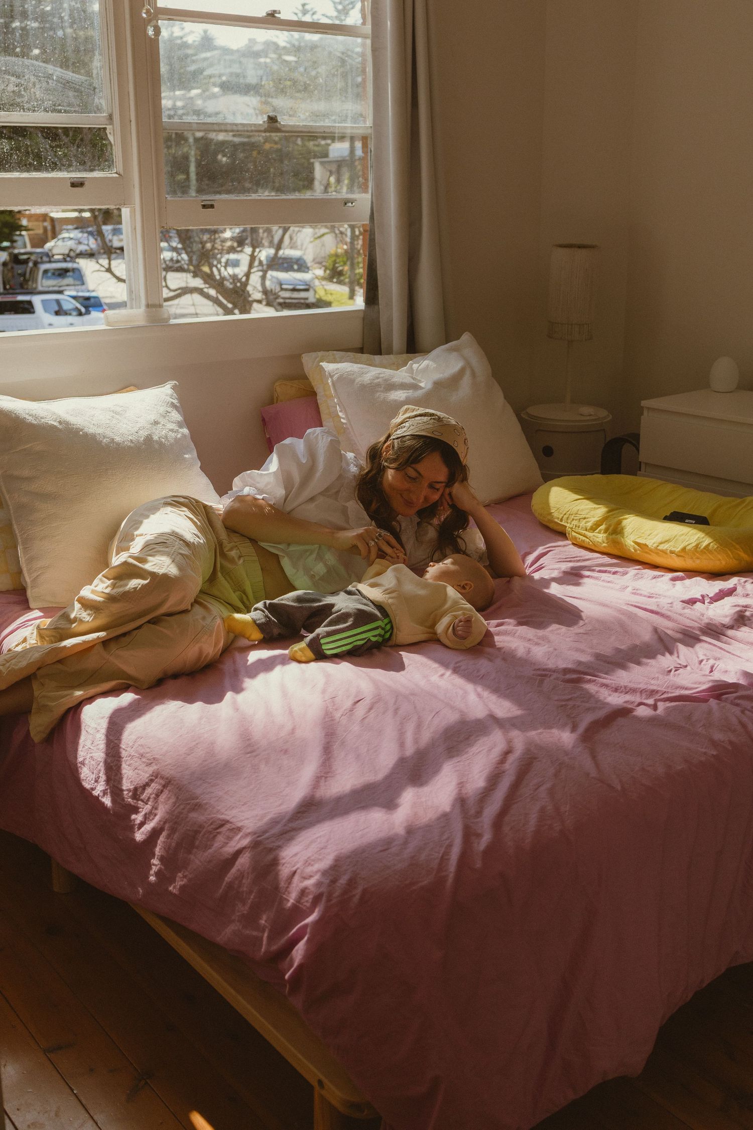 Young woman relaxing on pink bed with headphones, bathed in natural sunlight near a window.