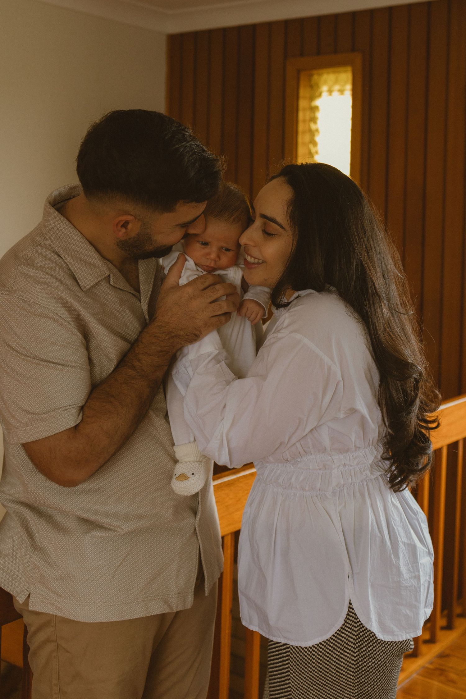 A couple sharing a tender moment kissing their newborn baby indoors near wooden railings.