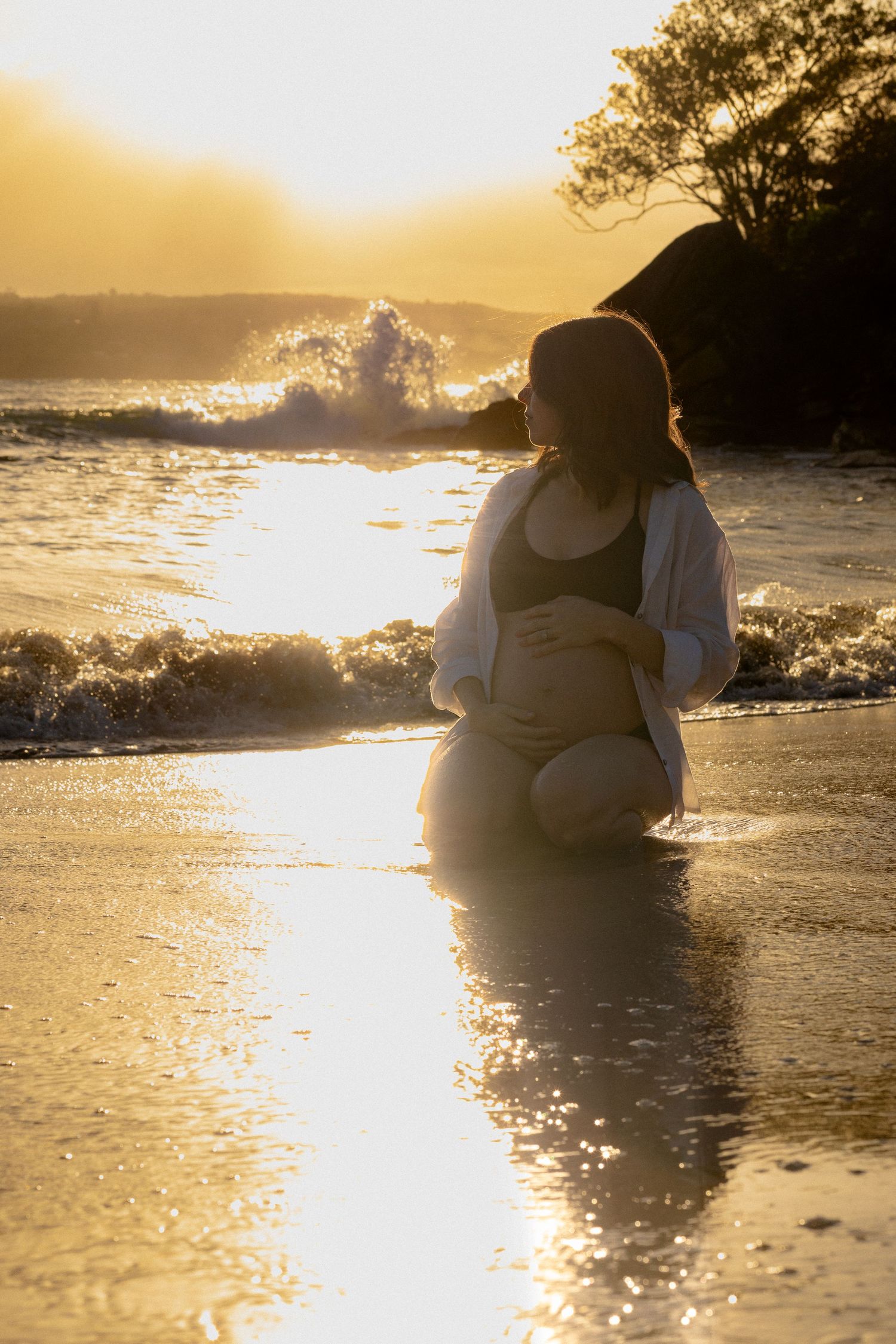 Pregnant woman kneeling on golden sunset beach, cradling her belly as ocean waves crash behind her.