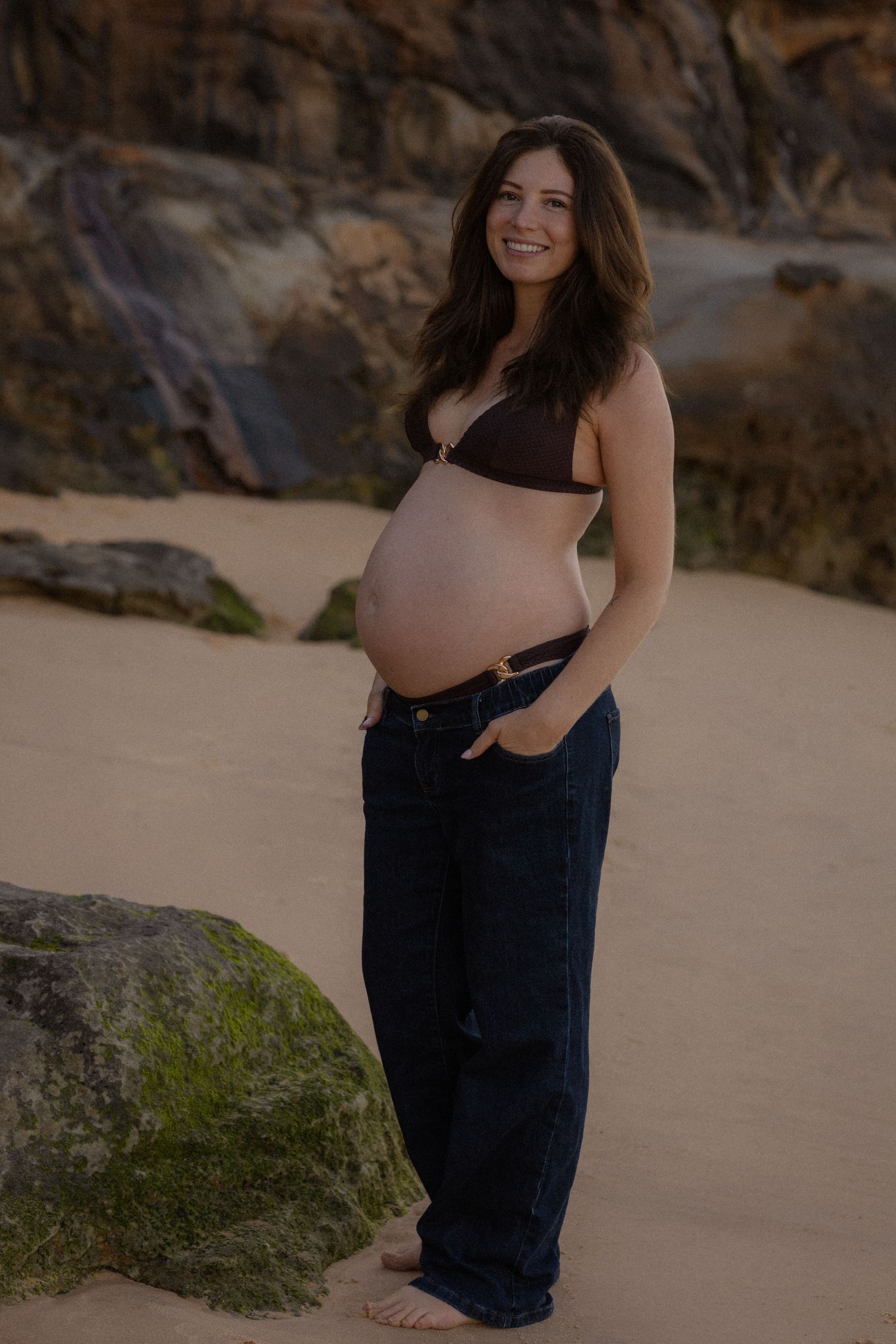 Smiling pregnant woman in bikini top and jeans posing on sandy beach with rocky backdrop.