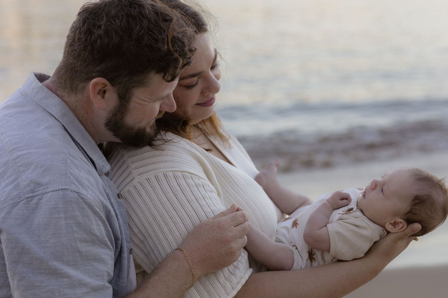 New parents tenderly admire their newborn baby by a serene beach at sunset.