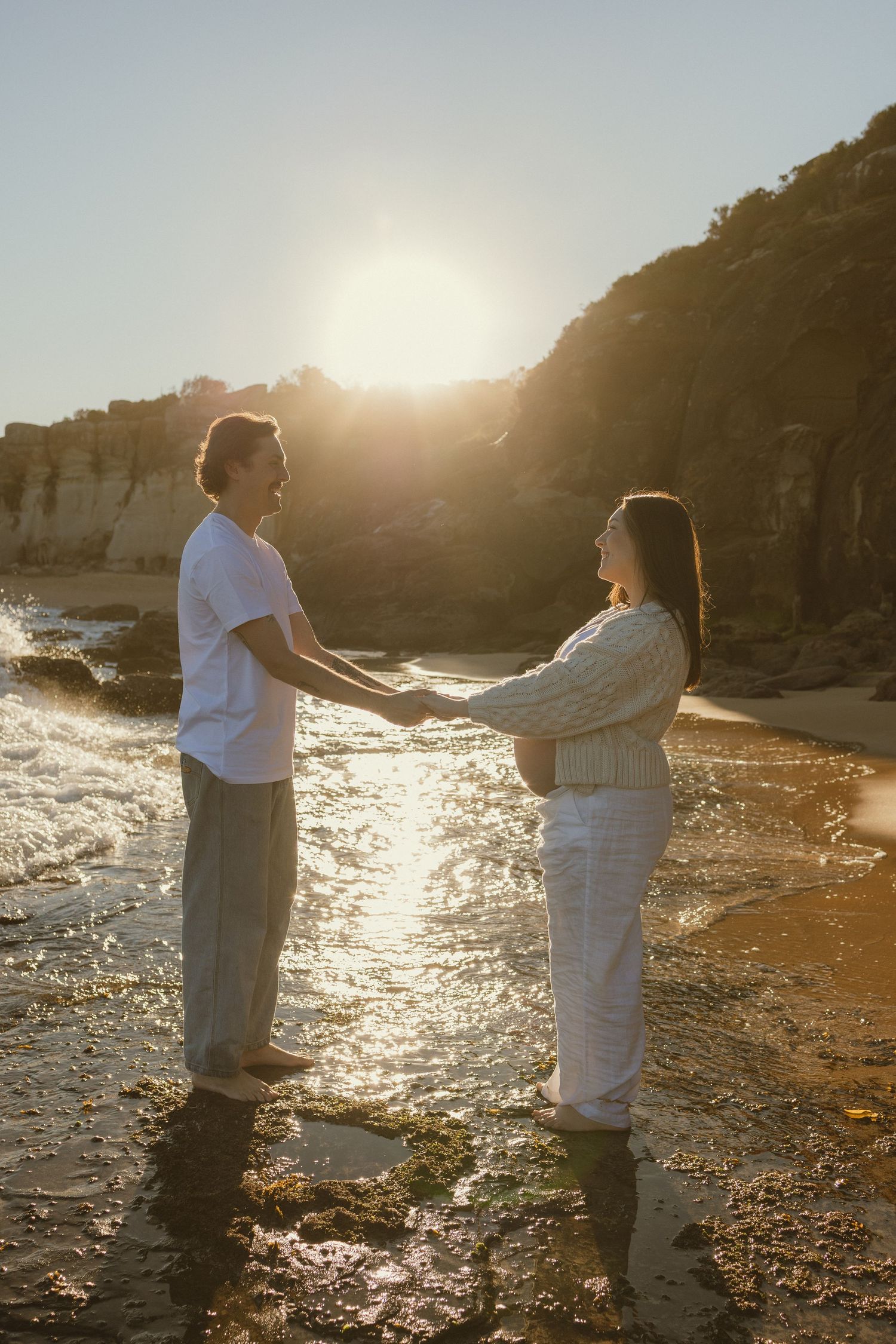 Couple holding hands on a sunlit beach with golden sunset light reflecting off the water near rocky cliffs.