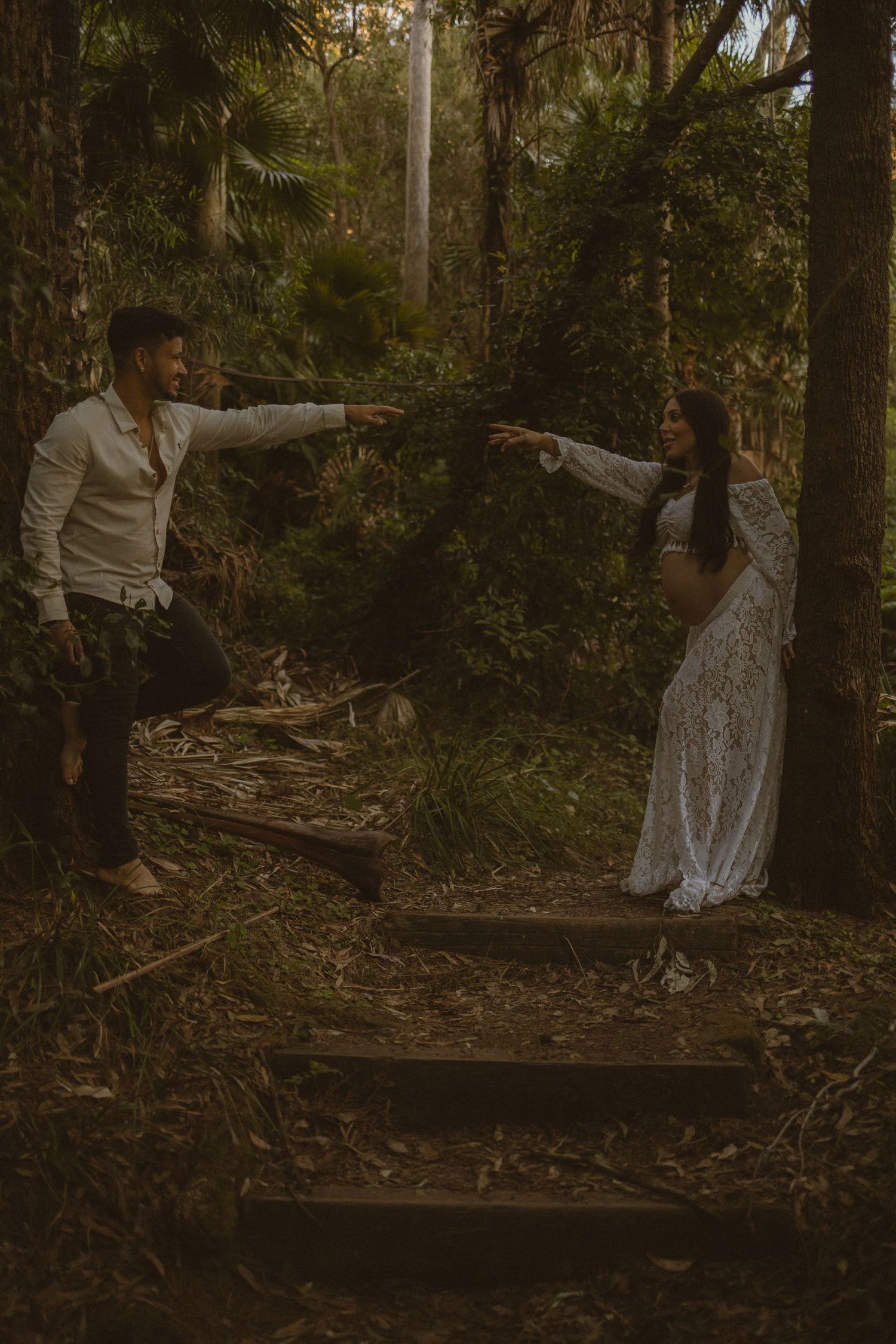 Pregnant woman in white lace dress reaches toward man in forest setting for a bohemian maternity photo shoot.