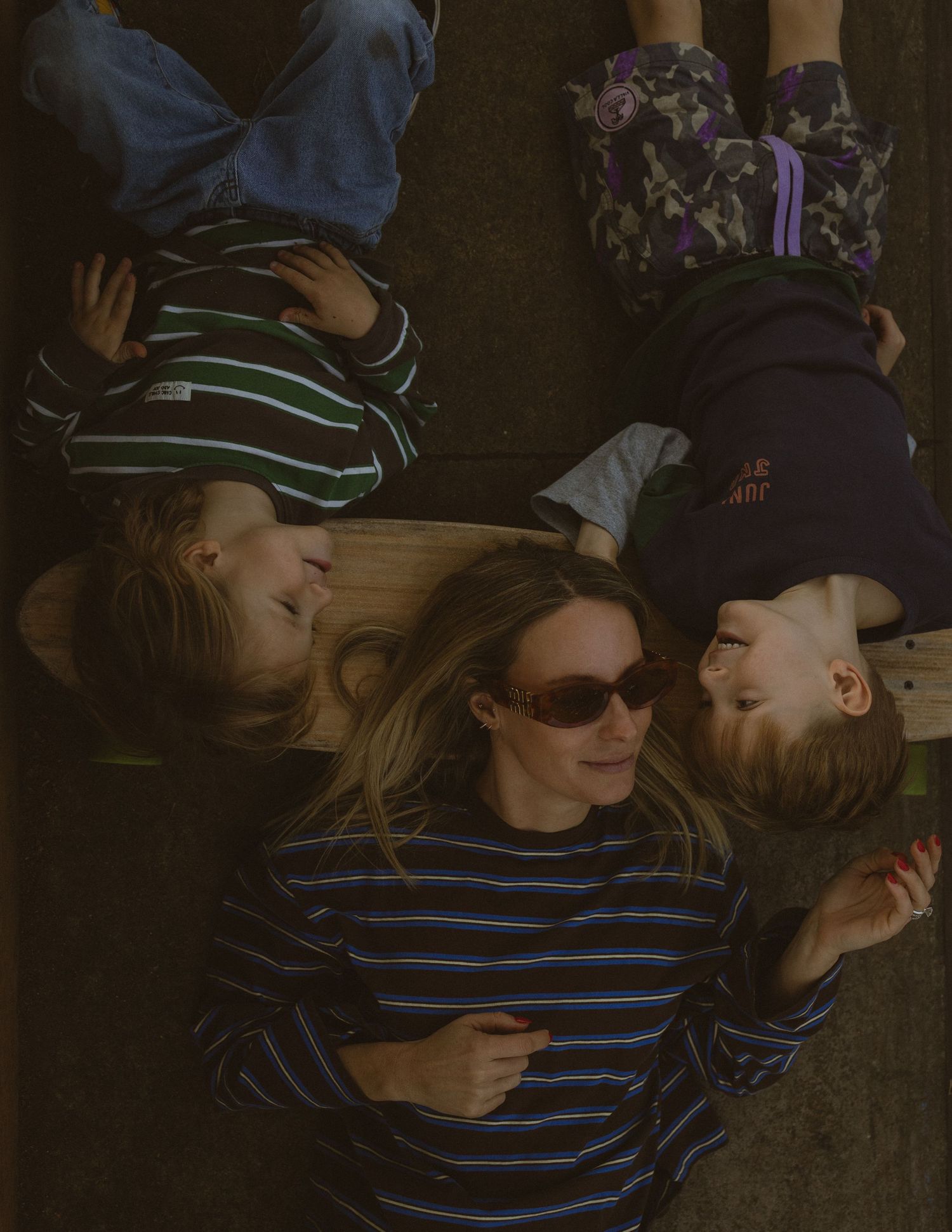 A woman wearing sunglasses lies on the ground smiling with two young children beside her outdoors.