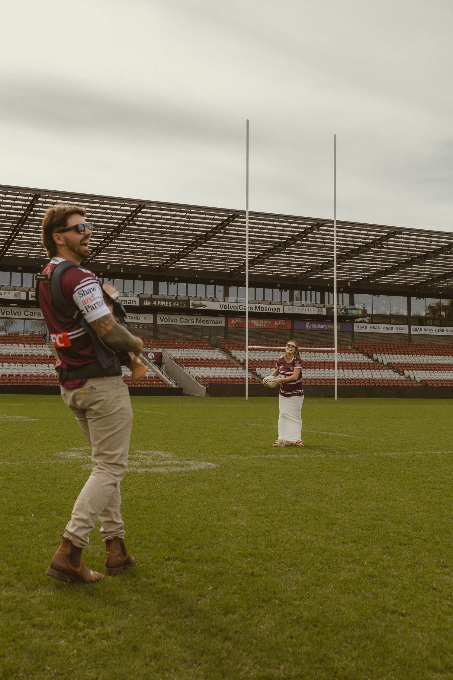 Couple in matching maroon jerseys playing on a rugby league field with goalposts and grandstand in background.
