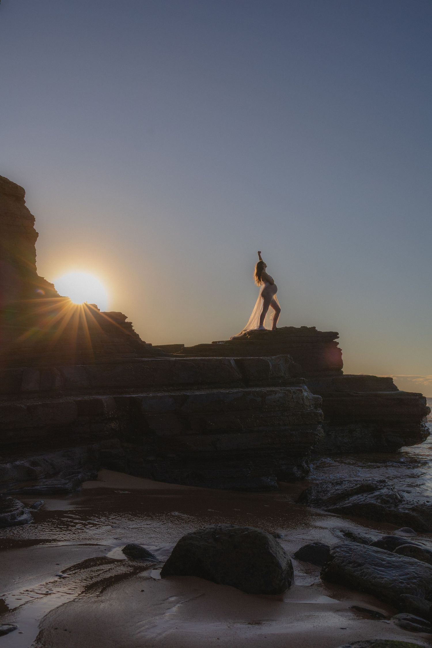 Silhouette of a woman in a flowing white dress posing on rocky coastal cliffs at golden sunset.