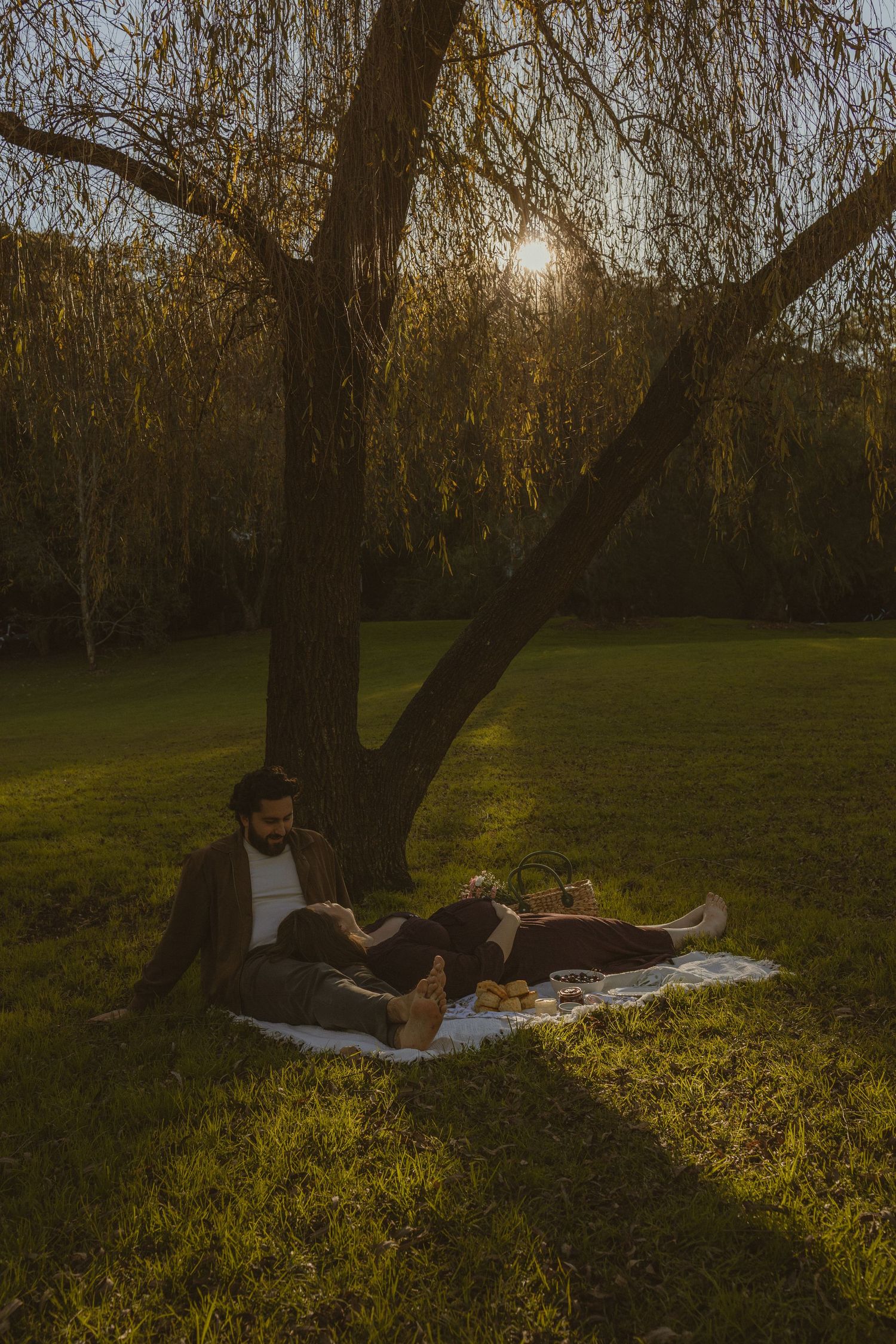 Couple relaxing on a blanket under a willow tree during golden hour sunset in a lush green park.