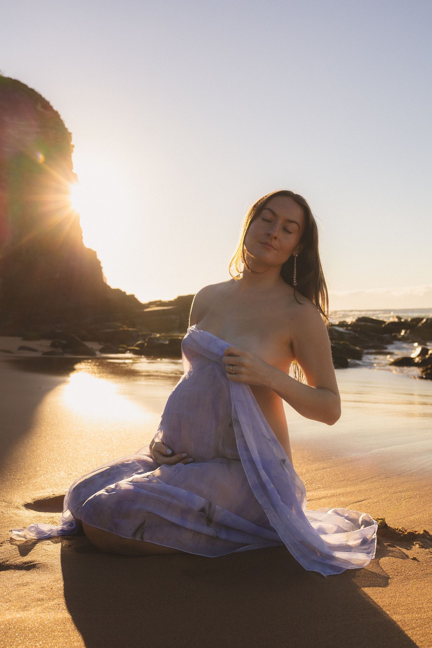 Pregnant woman draped in purple fabric sits on sandy beach at golden sunset with rocky cliffs.