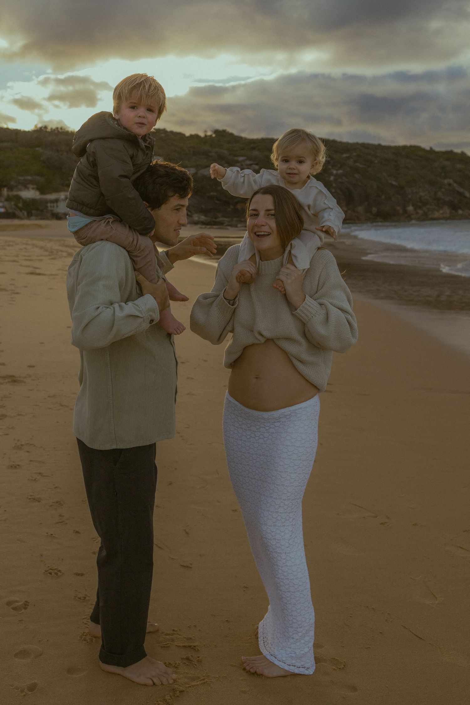 Family of four at golden hour beach, parents carrying toddlers on shoulders, smiling warmly on sandy shore.