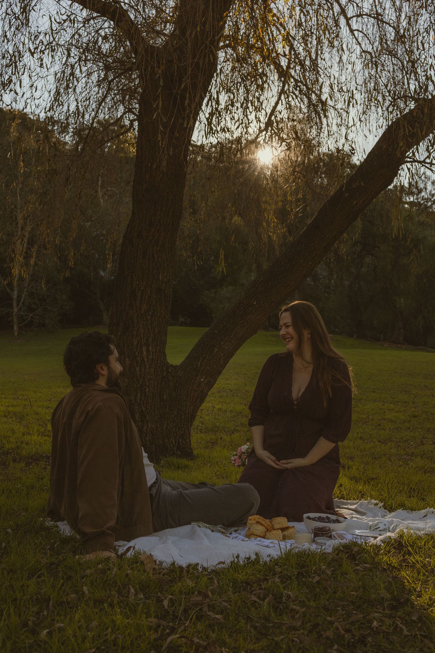 Couple enjoying a romantic sunset picnic on a blanket under a large tree in a lush green park.
